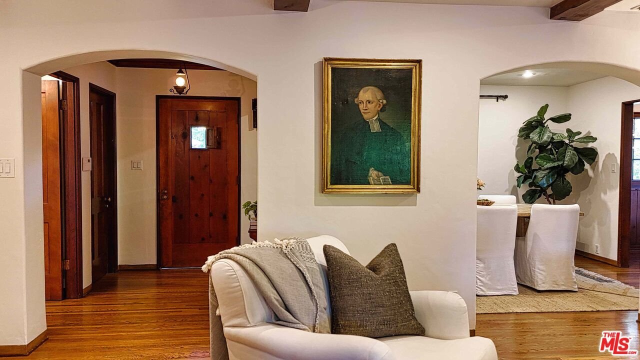 Living room with white walls, wooden floors, and archways. A white armchair with brown and beige cushions, a framed portrait of an older man on the wall, and an open dining area with a large green plant and white-covered chairs.