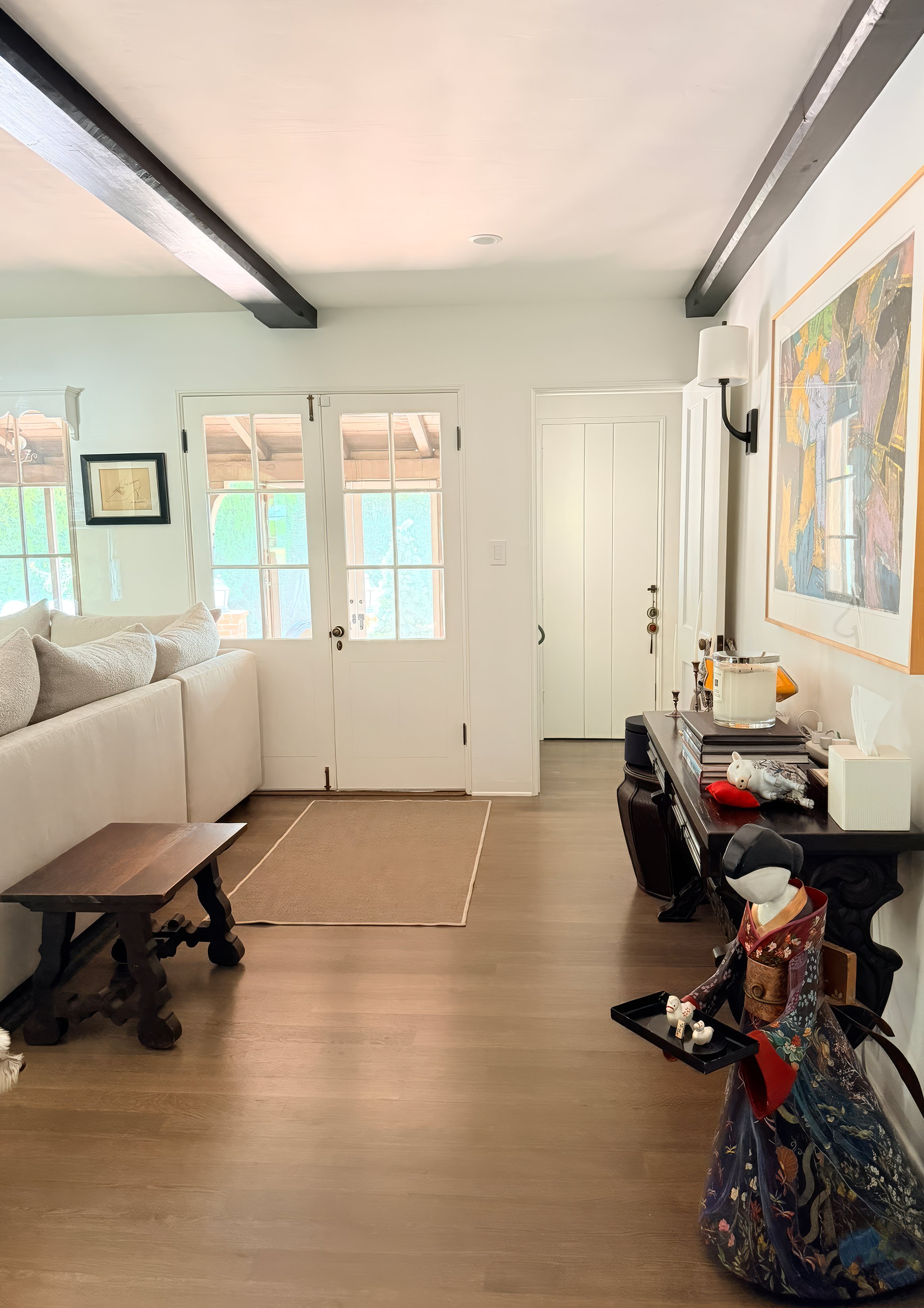 A living room with white walls, wooden flooring, and a large beige sectional sofa. Double glass doors lead to an outside porch. There is a black console table with Japanese dolls and decorative items. A large colorful abstract painting hangs on the w