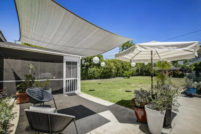 Backyard patio area with outdoor seating, large beige sunshade, white umbrella, potted plants, and string lights, overlooking a grassy lawn.