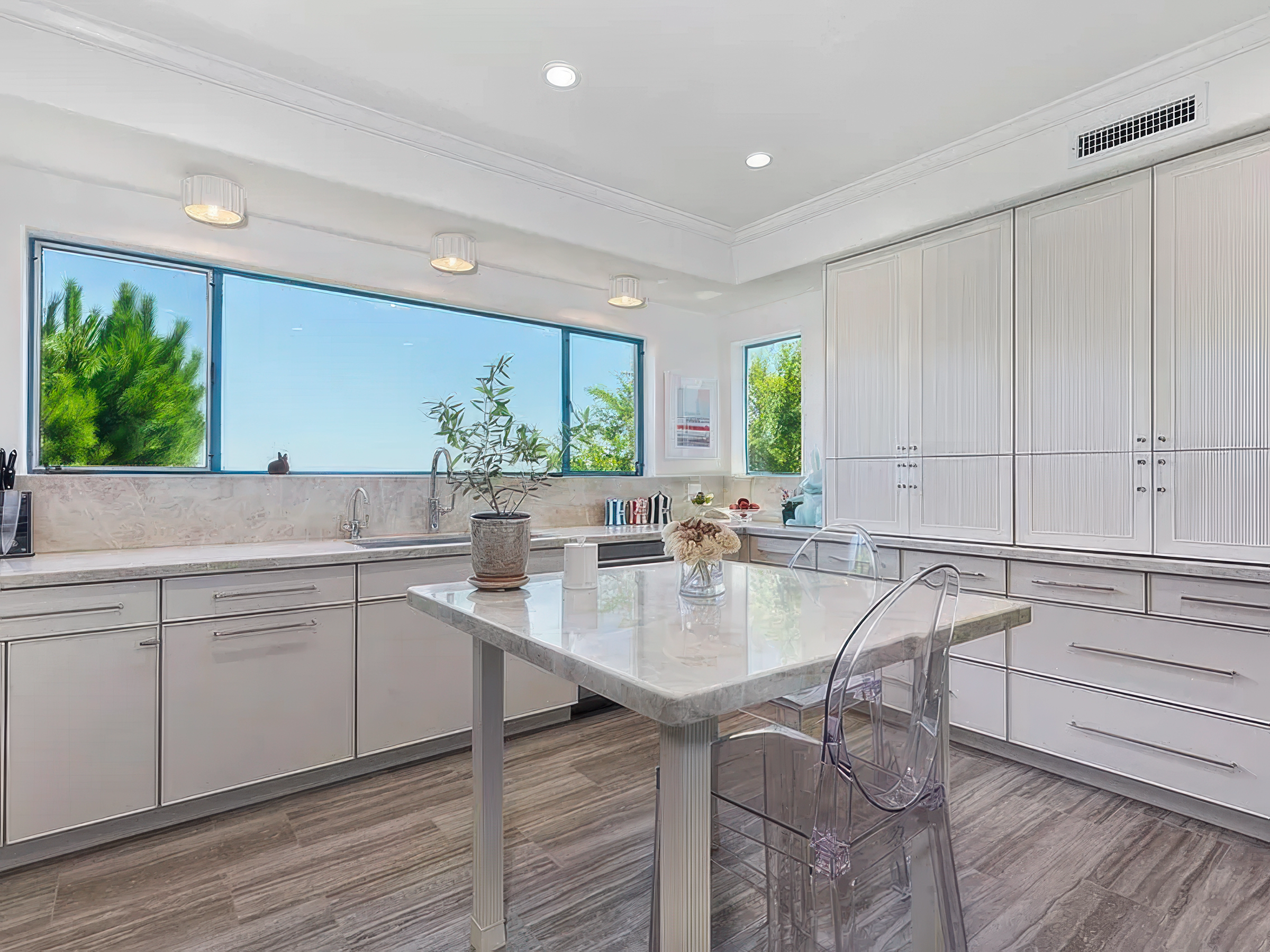 A white kitchen with large windows showing green trees outside, a marble countertop with a plant in a pot, a small figurine, and a kitchen island with a marble surface. There are white cabinets and clear plastic chairs around the island, with a hardw