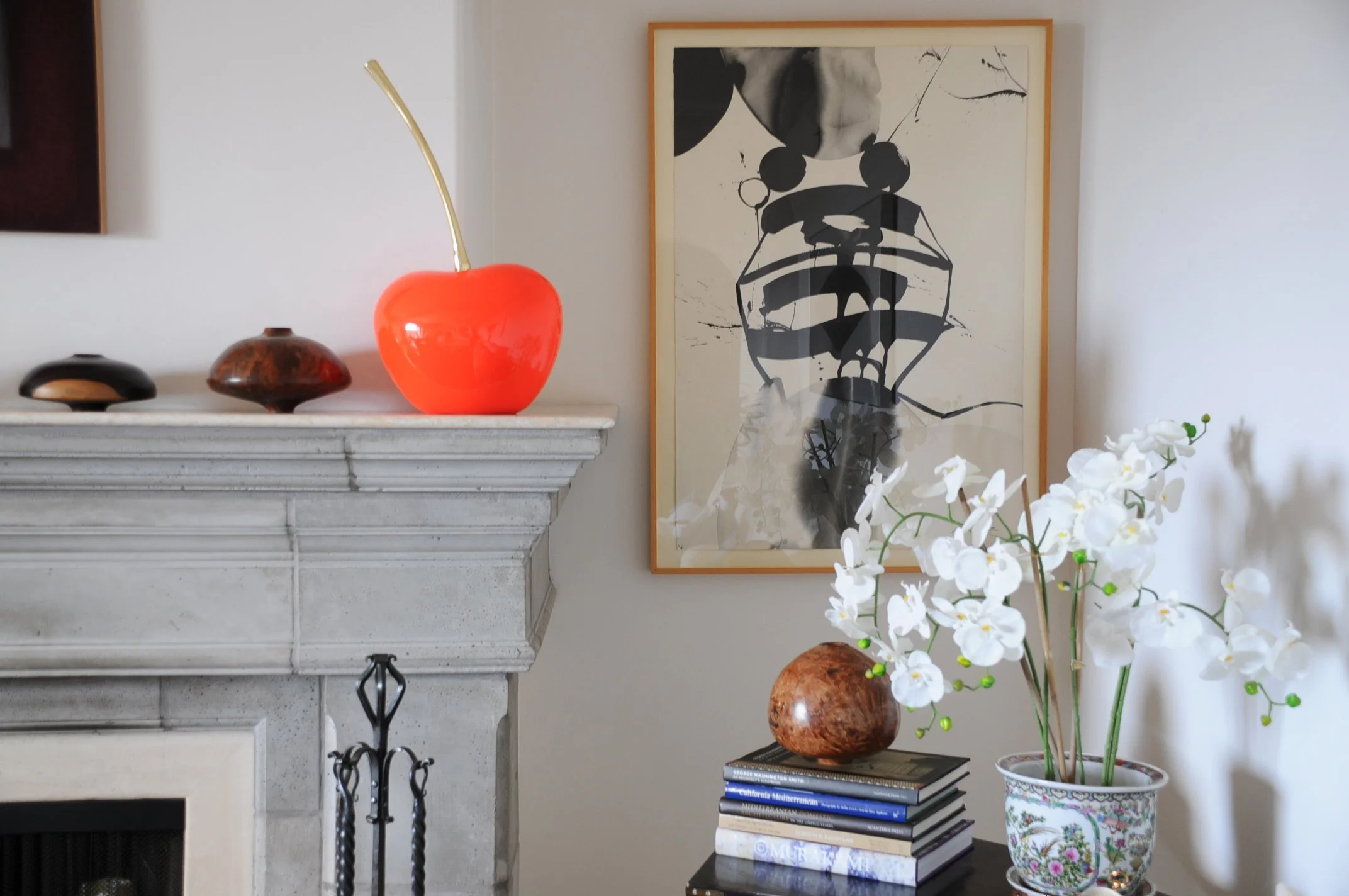 A living room corner with a white mantel, red ceramic cherry decorative object with a gold stem, two small wooden bowls, and a framed abstract black and white artwork on the wall. In front of the mantel, a stack of books with titles visible. To the r