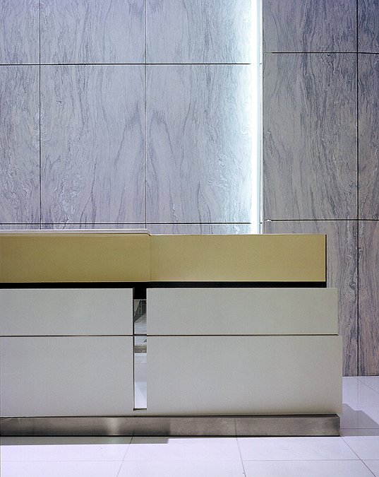 Modern reception desk with a light beige top, white and gray panels, set against a gray textured wall with vertical lighting.
