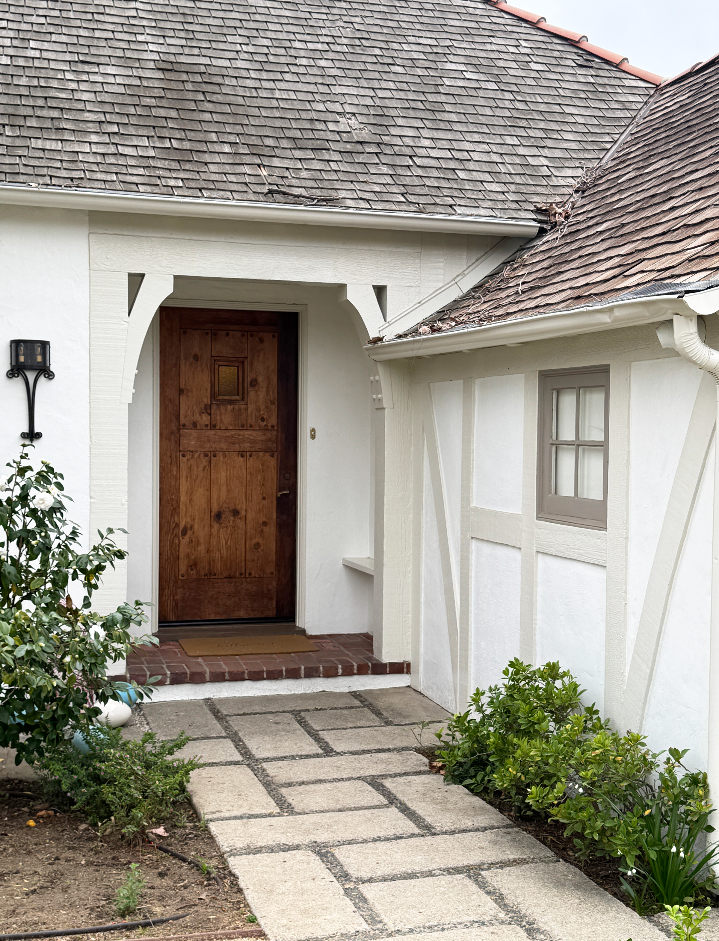 A house's front porch with a wooden door, white exterior walls, and a brick-stone walkway surrounded by green shrubs and plants.