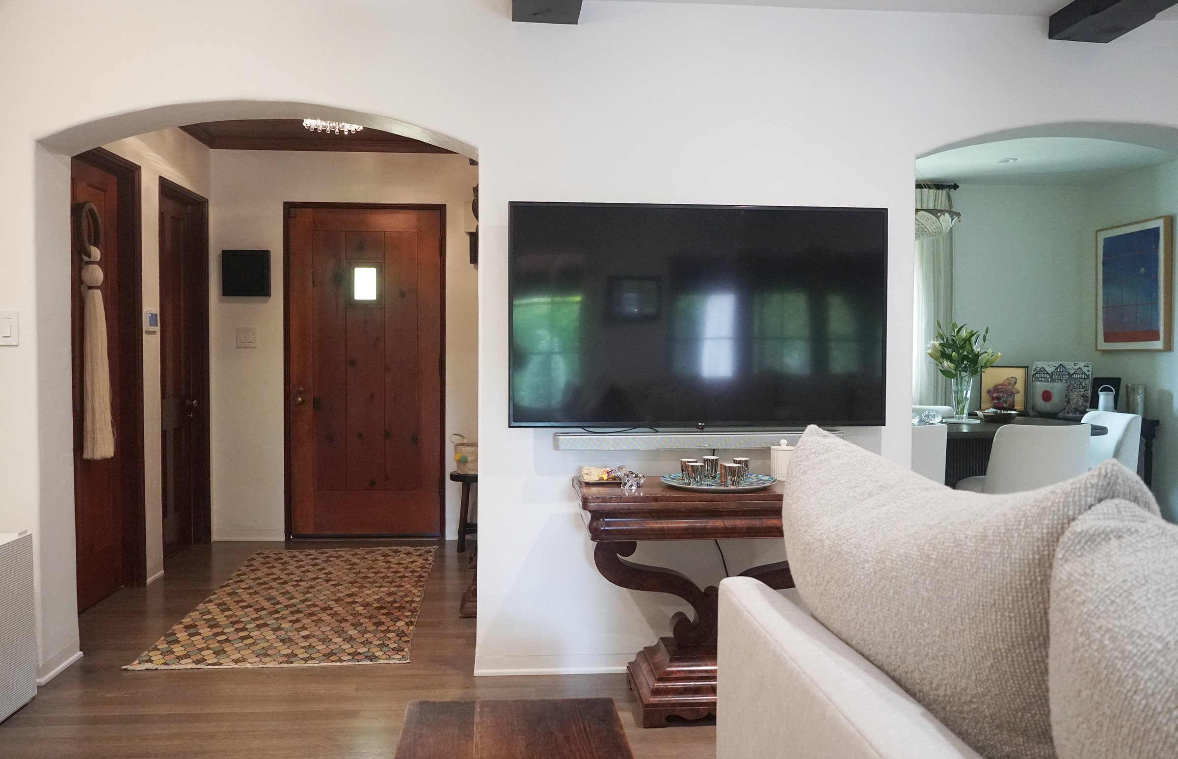 Living room with a beige sofa, wooden coffee table, flat-screen TV on a white wall, a side table with a vase of flowers, and a dining area with artwork on the wall.