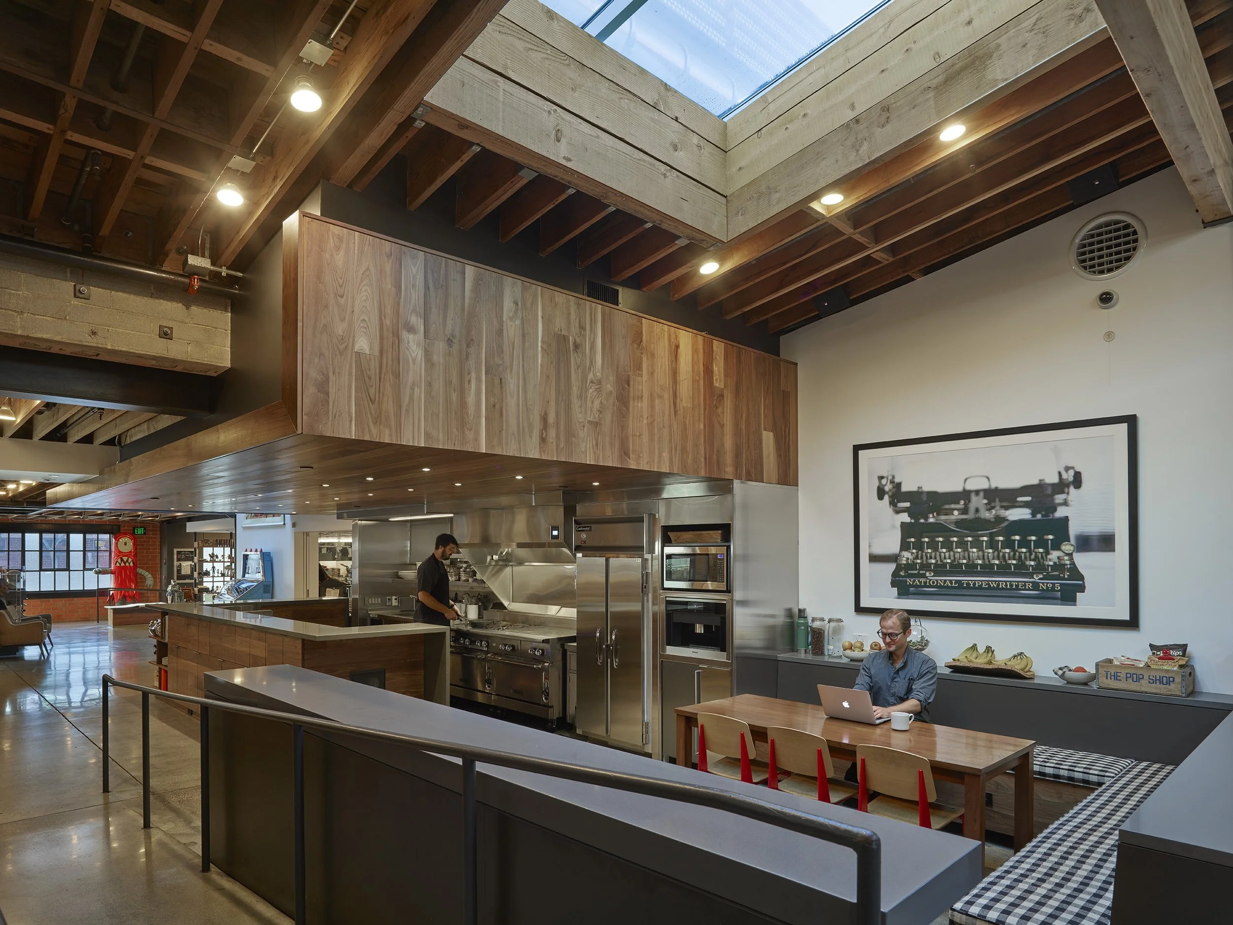 Interior of a modern cafe or coworking space with a wooden ceiling and overhead skylight, a man working on a laptop at a table, and decorative artwork on the wall.