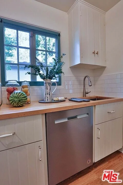Kitchen with a window, white cabinets, a wooden countertop, a small sink with a faucet, a dishwasher, and decorative jars and a watermelon on the counter.