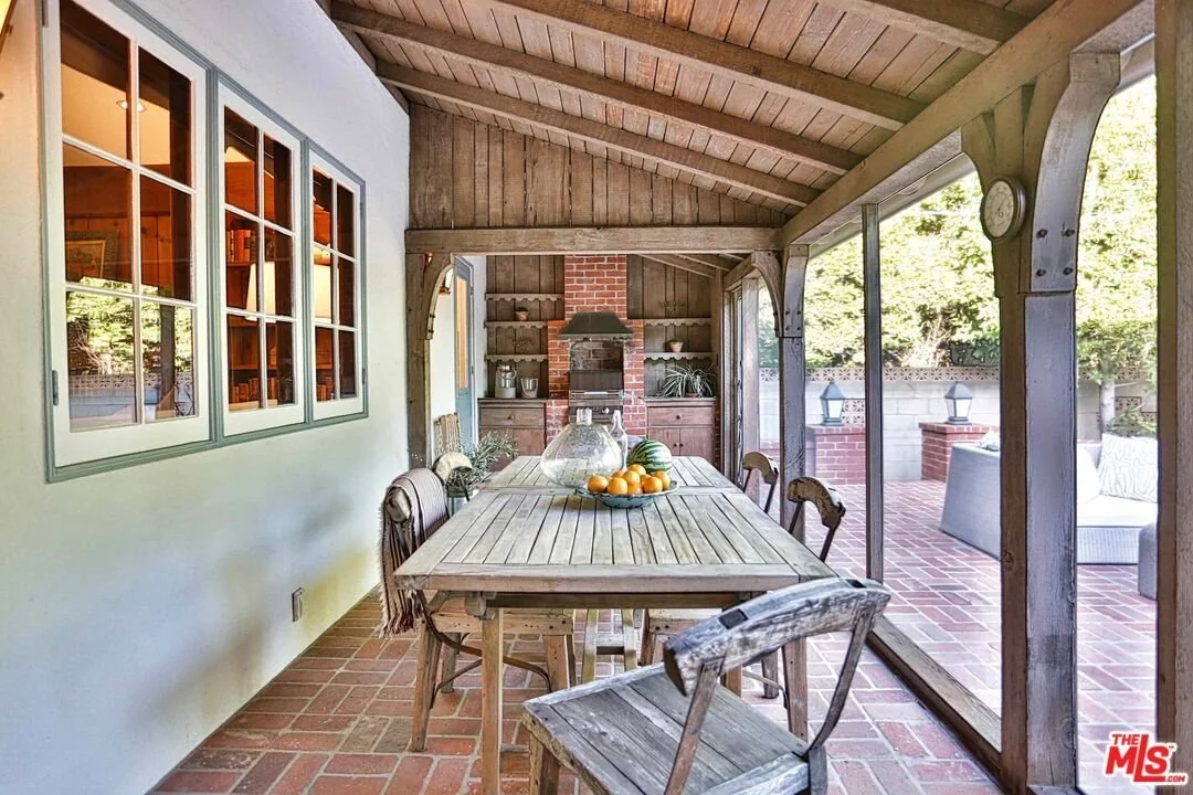 Enclosed outdoor patio with wooden ceiling, brick accents, and a rustic dining table with chairs, set with a fruit bowl and glass decanter, overlooking a brick fireplace and seating area outside.