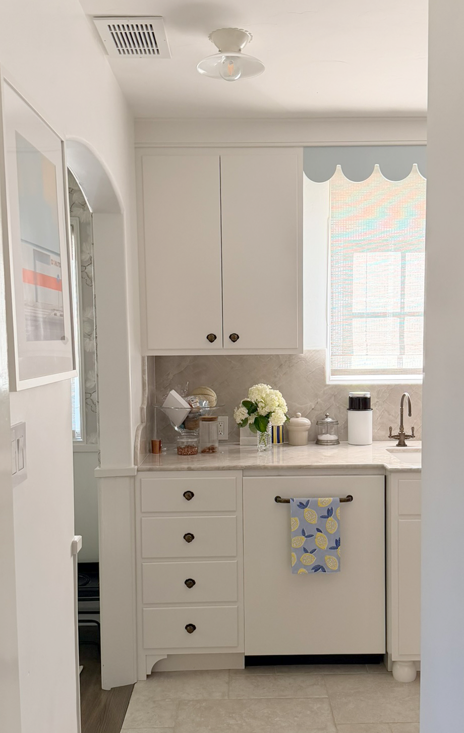 A small kitchen with white cabinets and a marble countertop, decorated with a vase of white flowers, jars, and a lemon-patterned towel hanging on a bar.