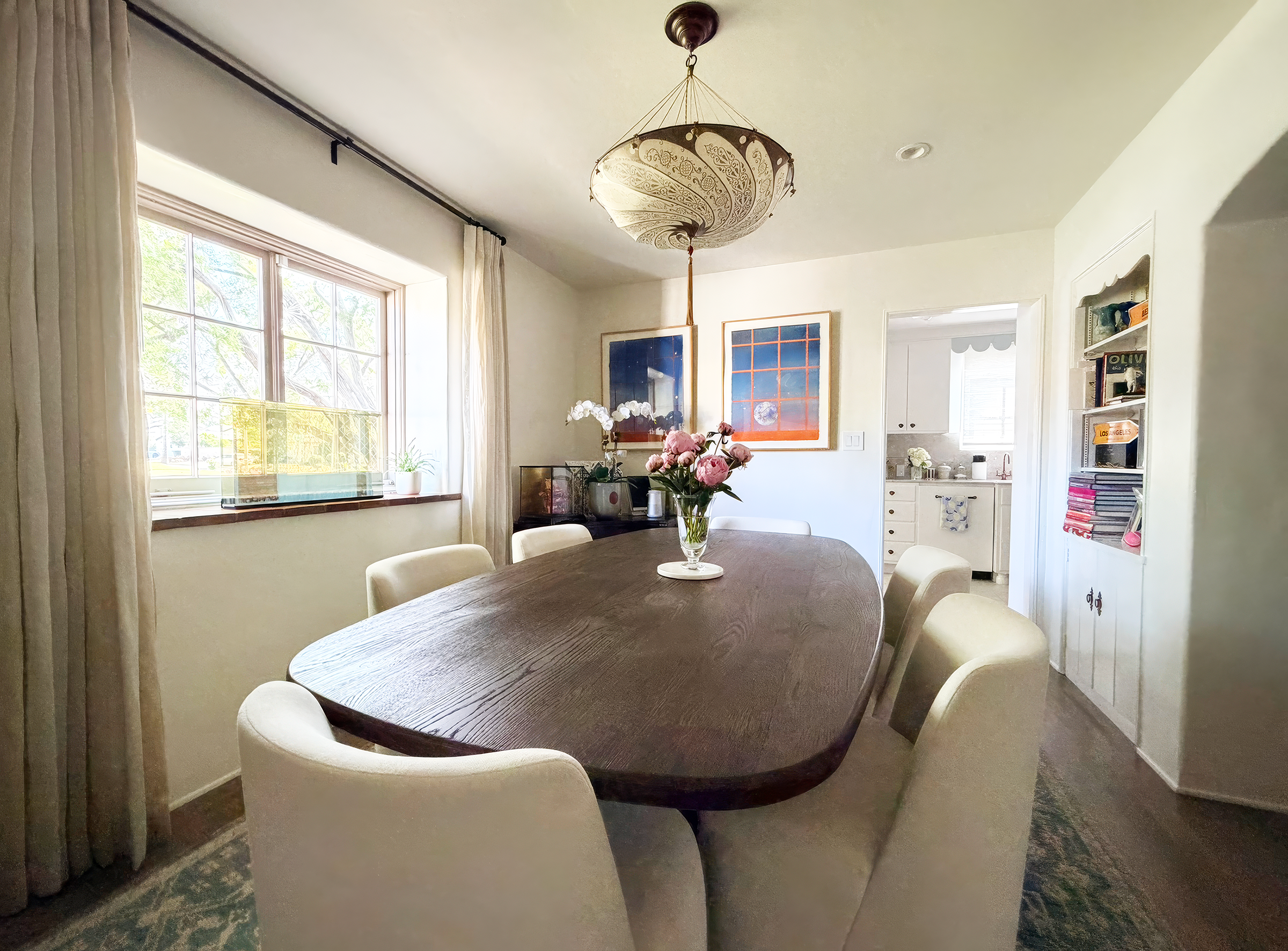 Interior of a dining room with a wooden oval table surrounded by eight cream-colored chairs, a vase of pink roses, large windows with curtains, wall art, and a chandelier hanging from the ceiling.