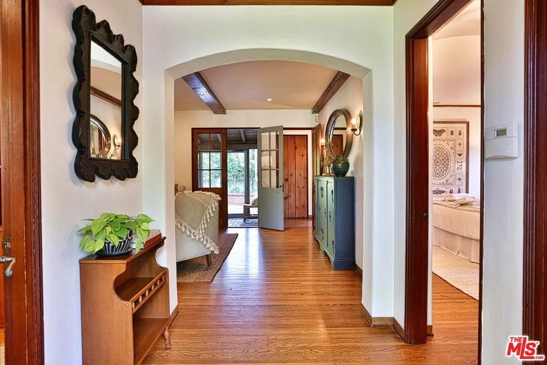 Interior view of a cozy home hallway with wooden floors, decorative mirrors, potted plant, and view into living room and bedroom.