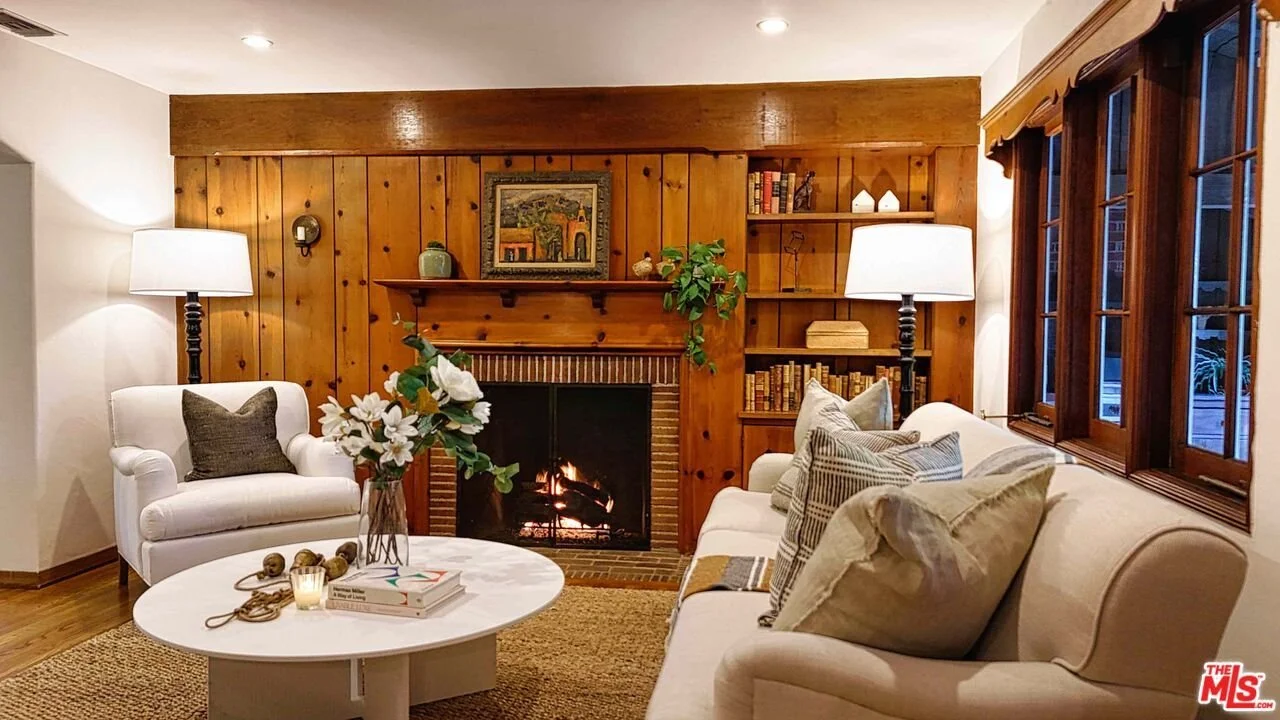Living room with a fireplace, a white armchair, and a white sofa with decorative pillows. There are two floor lamps, a round coffee table with flowers, books, and a candle. The wood-paneled wall behind the fireplace has a painting, some bookshelves, 