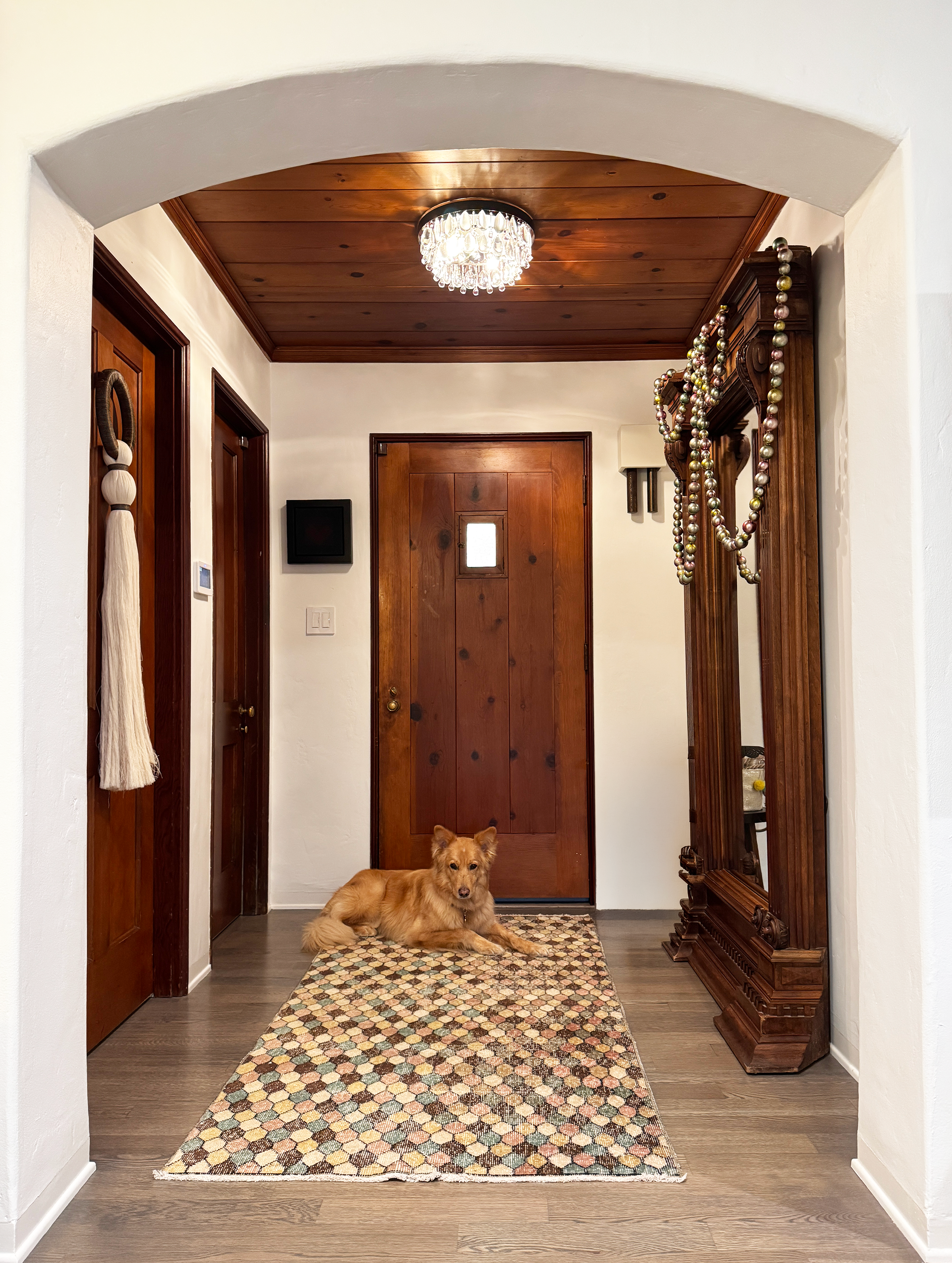 Interior view of a hallway with a wooden ceiling, decorative chandelier, a dog lying on a patterned rug near a wooden door, and a large ornate wooden mirror with bead garland decorations hanging.