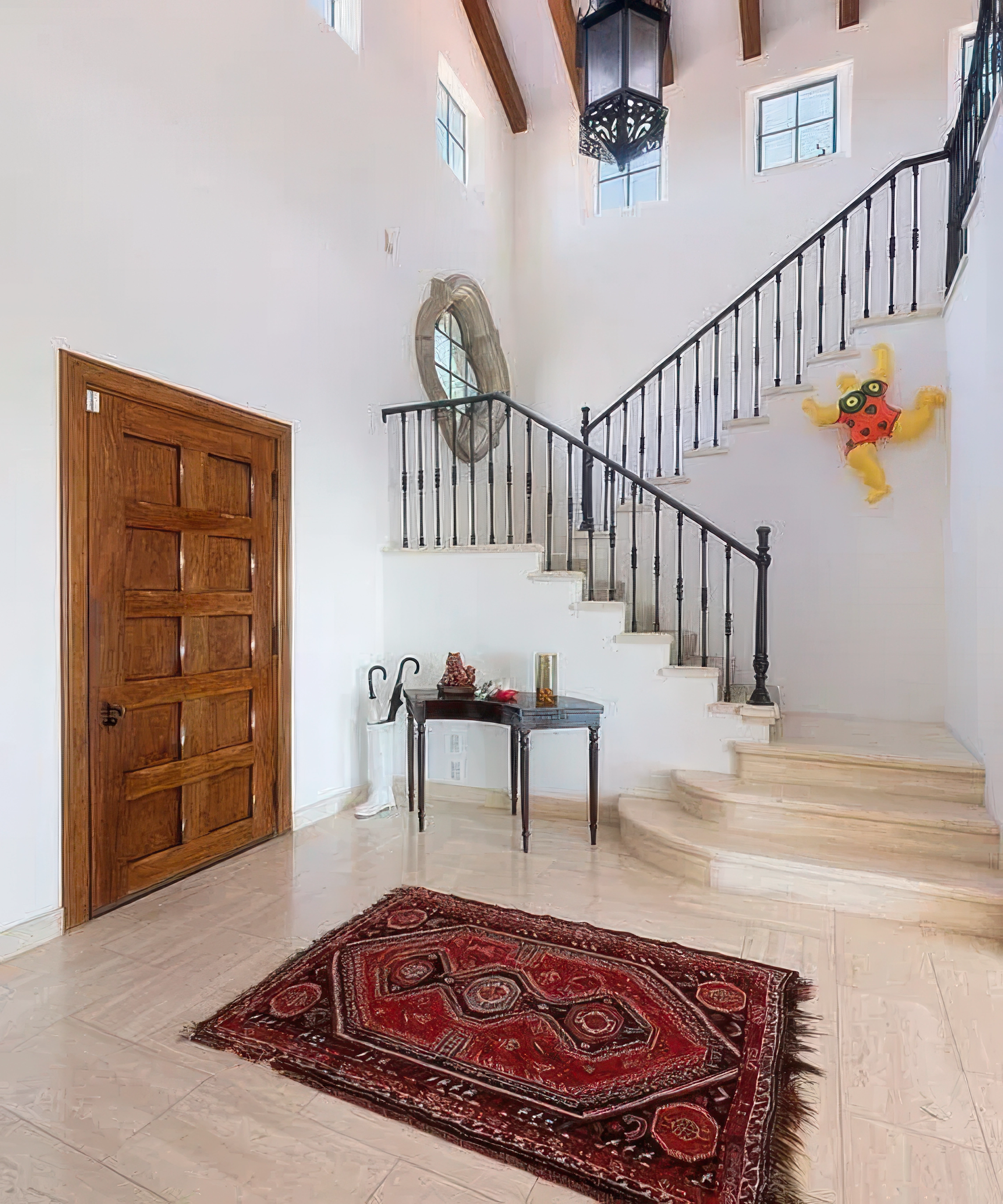 An entryway with a wooden door, a patterned red rug on a glossy cream-colored tile floor, a staircase with white steps and a black metal railing, small rectangular windows, a mirror or window round frame on the wall, a small table with decorations, a