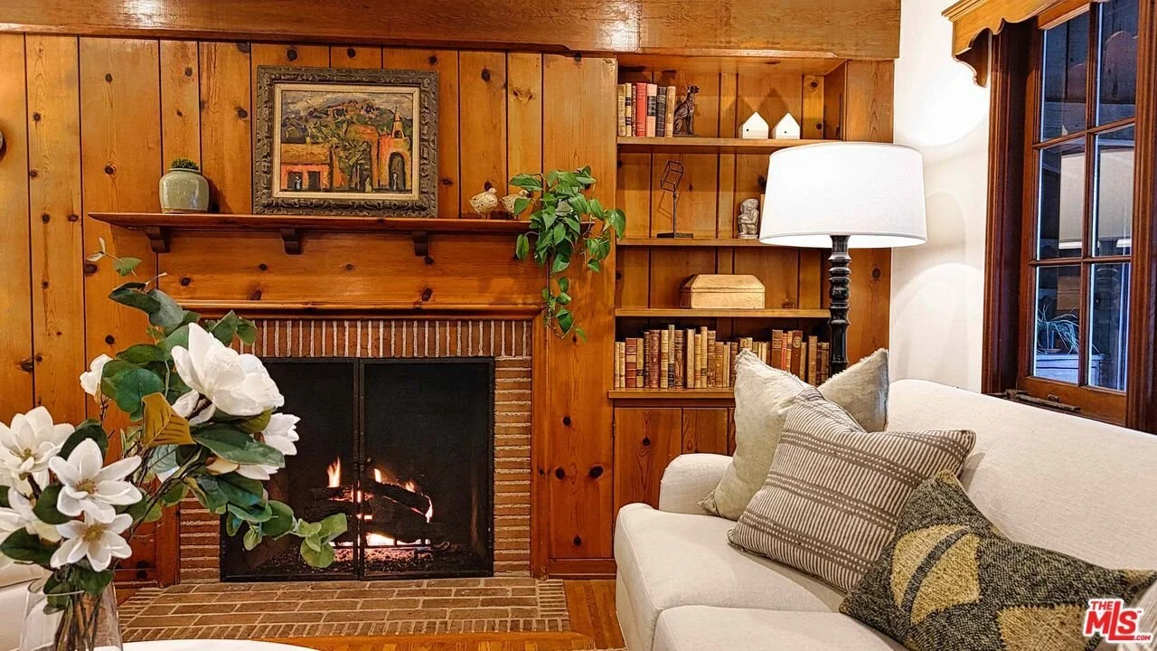 Living room with a brick fireplace, wood-paneled walls, a white sofa with decorative pillows, a lamp, a window, and built-in bookshelves with books and decorative items.