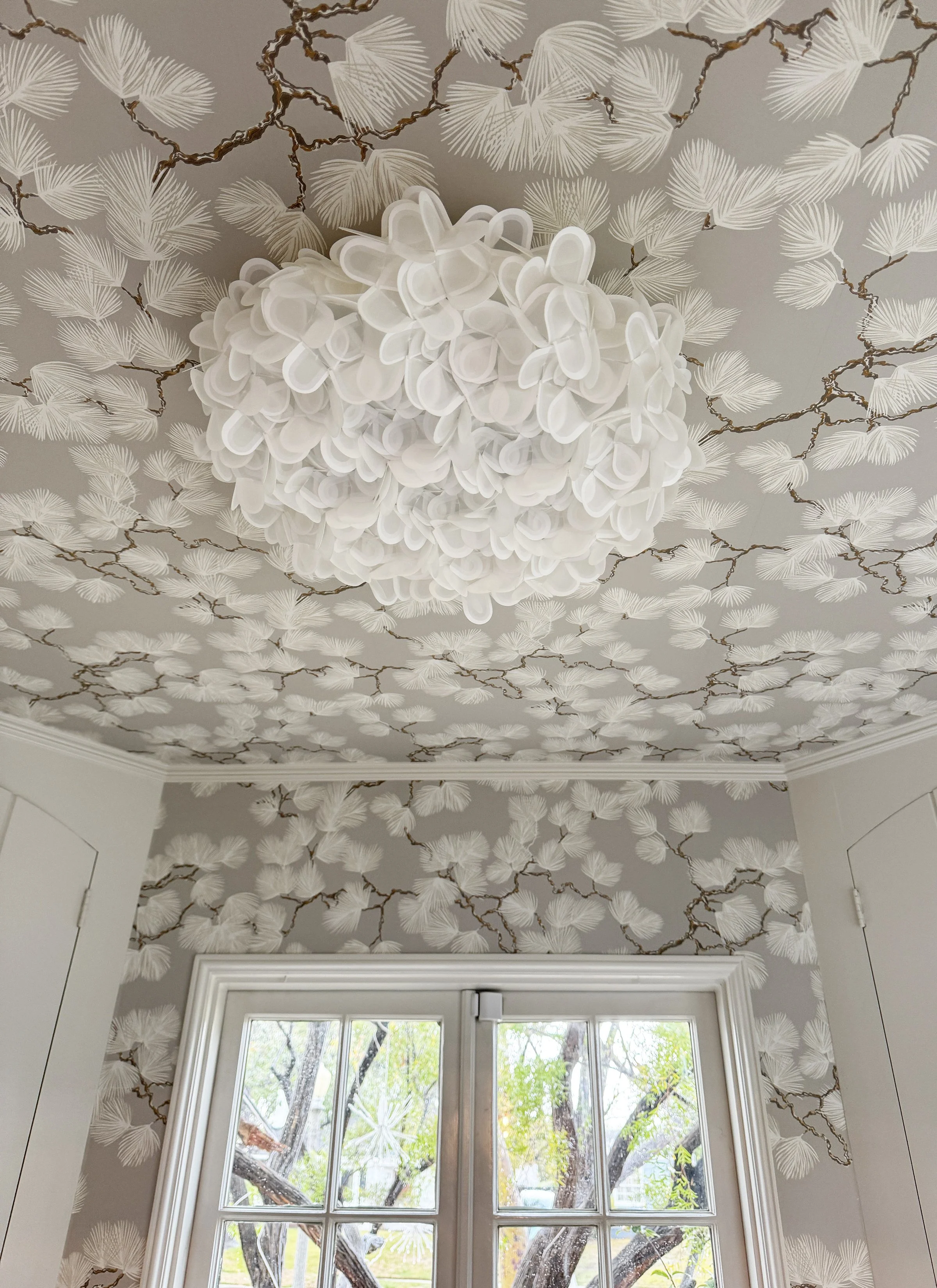 A view from inside a room looking up at a ceiling with floral wallpaper and a modern white pendant light fixture. The room has a large window with multiple panes, showing trees and greenery outside.