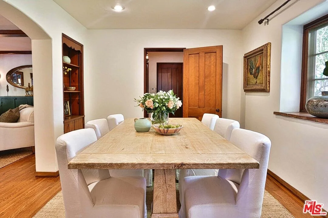 A dining room with a wooden table, eight white upholstered chairs, a vase with pink and white flowers, a small green bowl, and a decorative plate. The room has two windows with wooden frames and a framed painting of a butterfly on the wall.