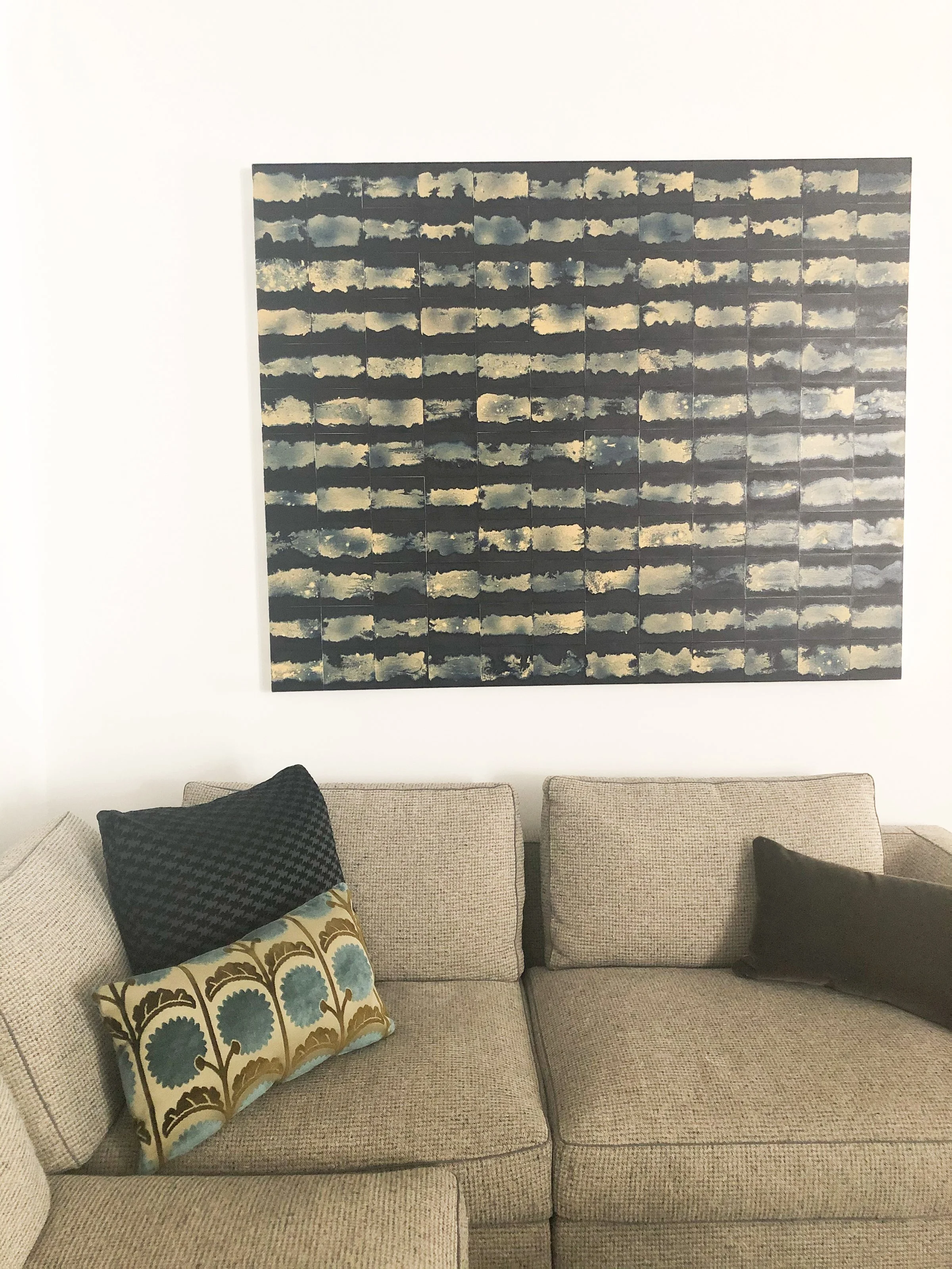 A beige sectional sofa with three decorative pillows, one patterned with blue, green, and gold, and two solid black pillows, against a white wall with a large abstract painting of clouds.