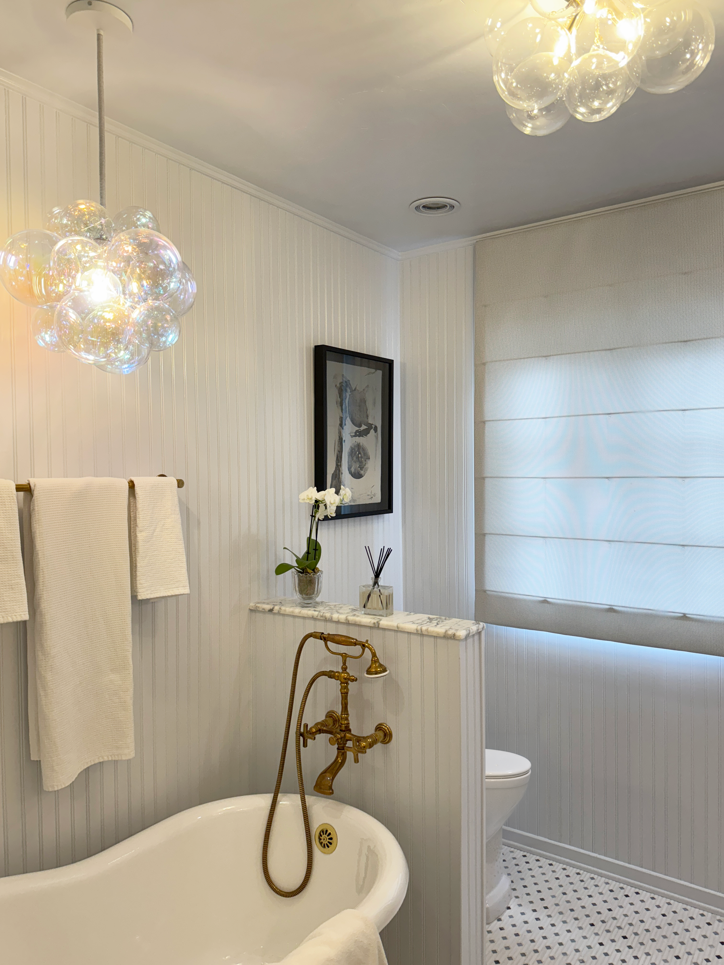 A vintage-style bathroom with a clawfoot bathtub, antique brass fixtures, a modern toilet, and decor including framed art, a potted orchid, and patterned window blinds.