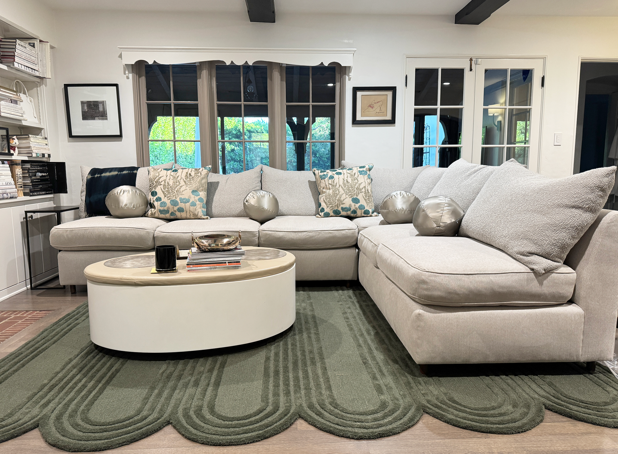 Living room with a large light gray sectional sofa decorated with patterned and metallic pillows, a white oval coffee table with magazines and decor, a green rug with concentric circle pattern, and large bay windows with a view of greenery outside.