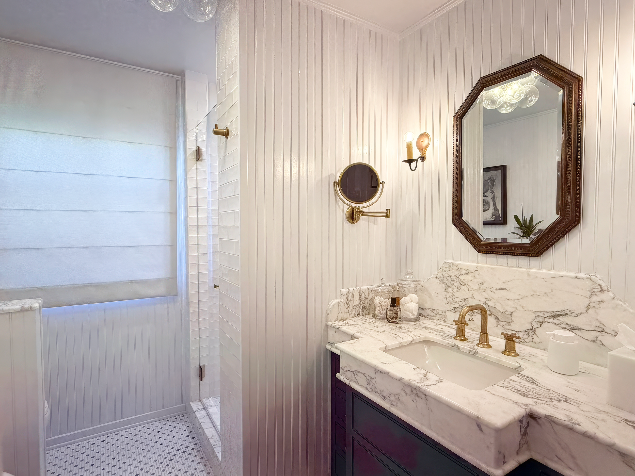 A bathroom with a marble countertop, a gold faucet, a large mirror with a carved wooden frame, a small round mirror, and wall-mounted light fixtures, with a shower area visible on the left side.