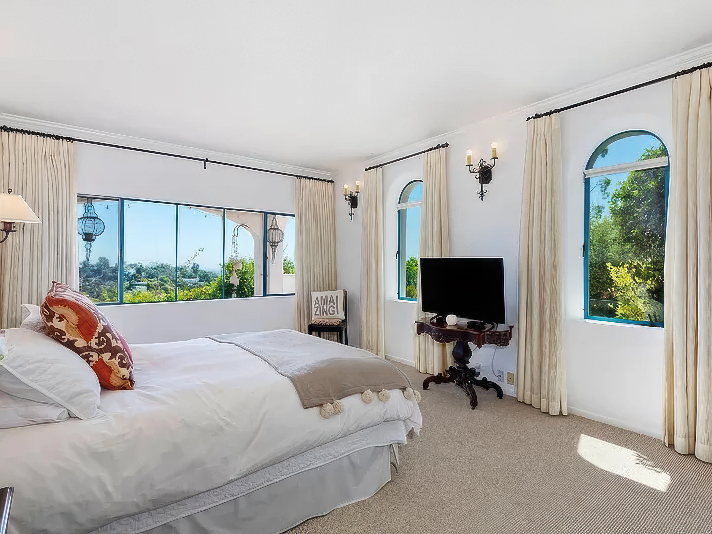 Bright bedroom with white curtains, arched windows showing green trees outside, and a bed with white bedding and patterned pillows. A small table with a television stand, a lamp, and decorative wall sconces with candles are visible.