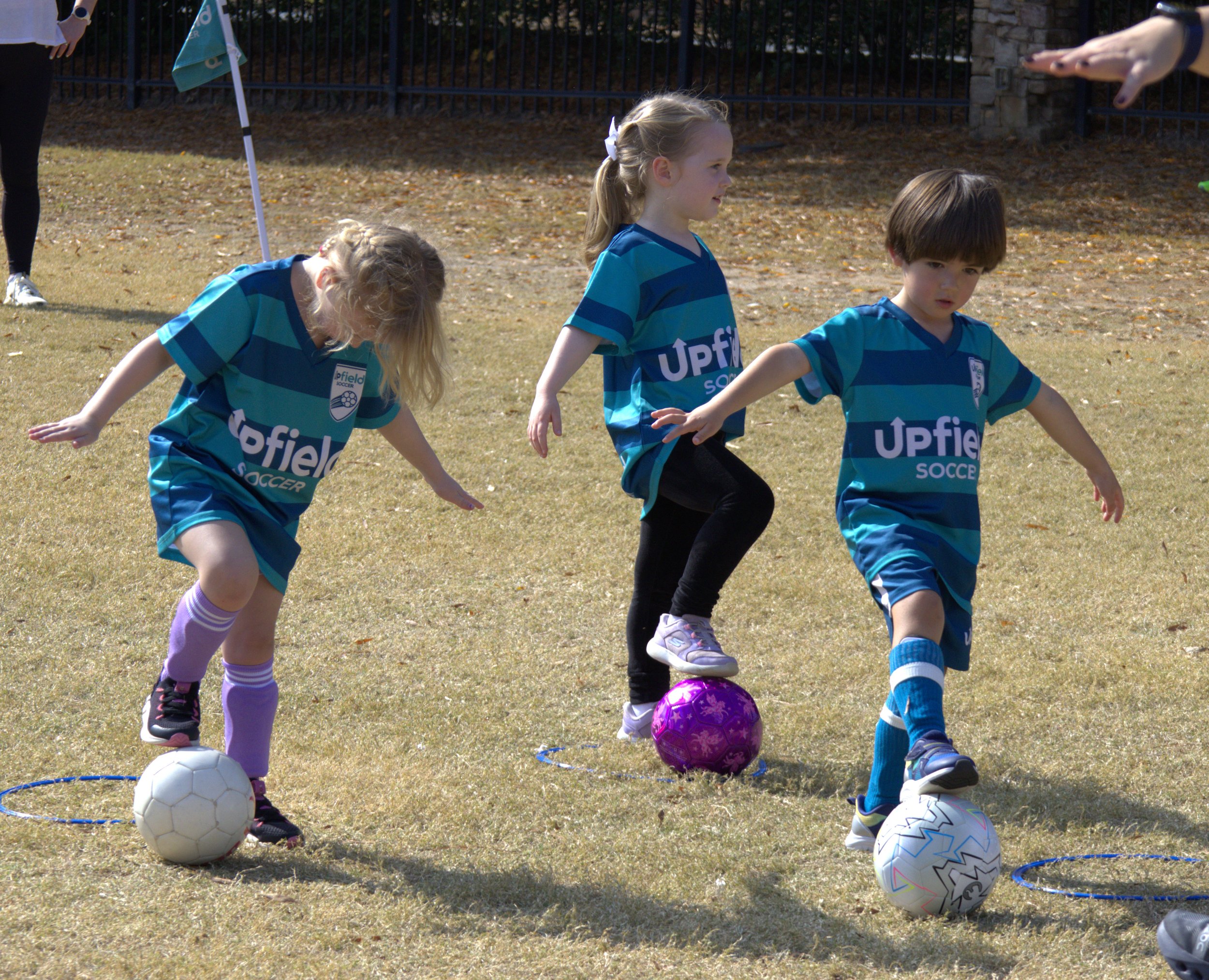 three kids with soccer balls balance with one foot on top of their ball