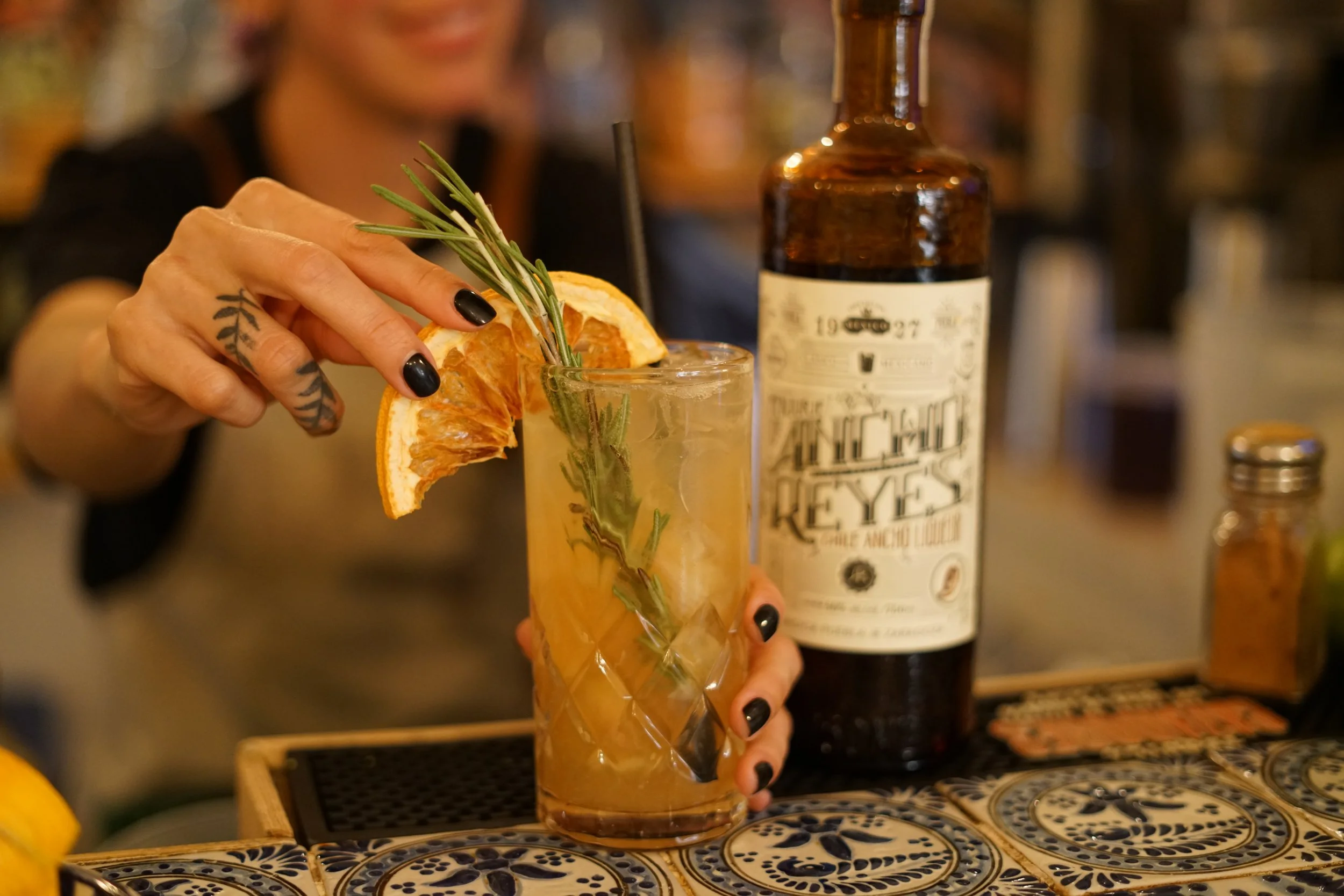 Bartender garnishing cocktail with rosemary and dried orange slice, bottle of Ancho Reyes in background.