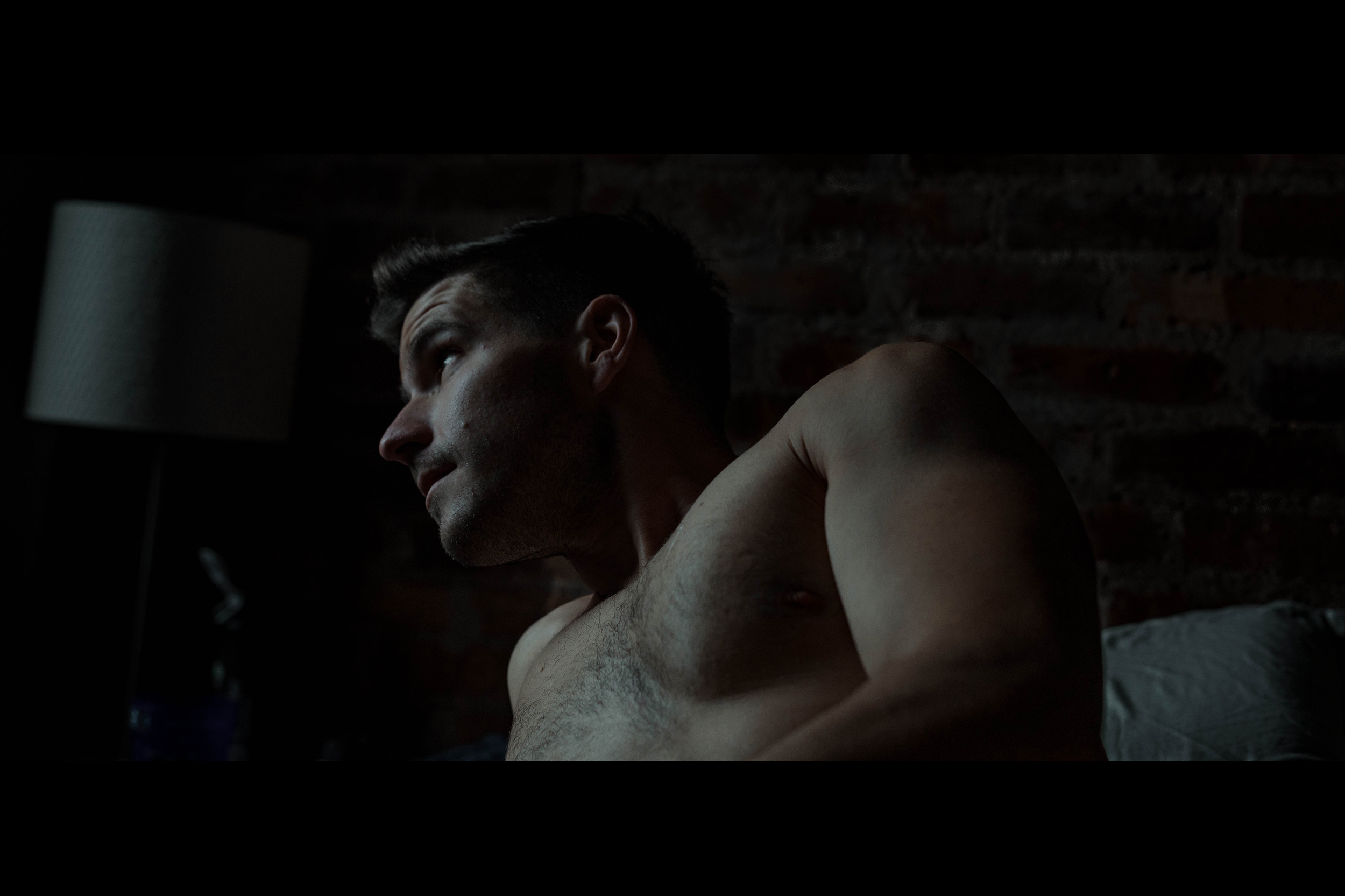 Cinematic portrait of shirtless model in dark bedroom looking out the window