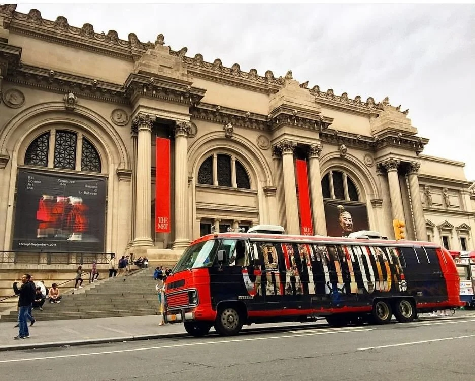 A tour bus with a red, blue, and black design Gratitude Bus parked in front of a large, historic building with columns and arched windows, with people walking on the sidewalk.