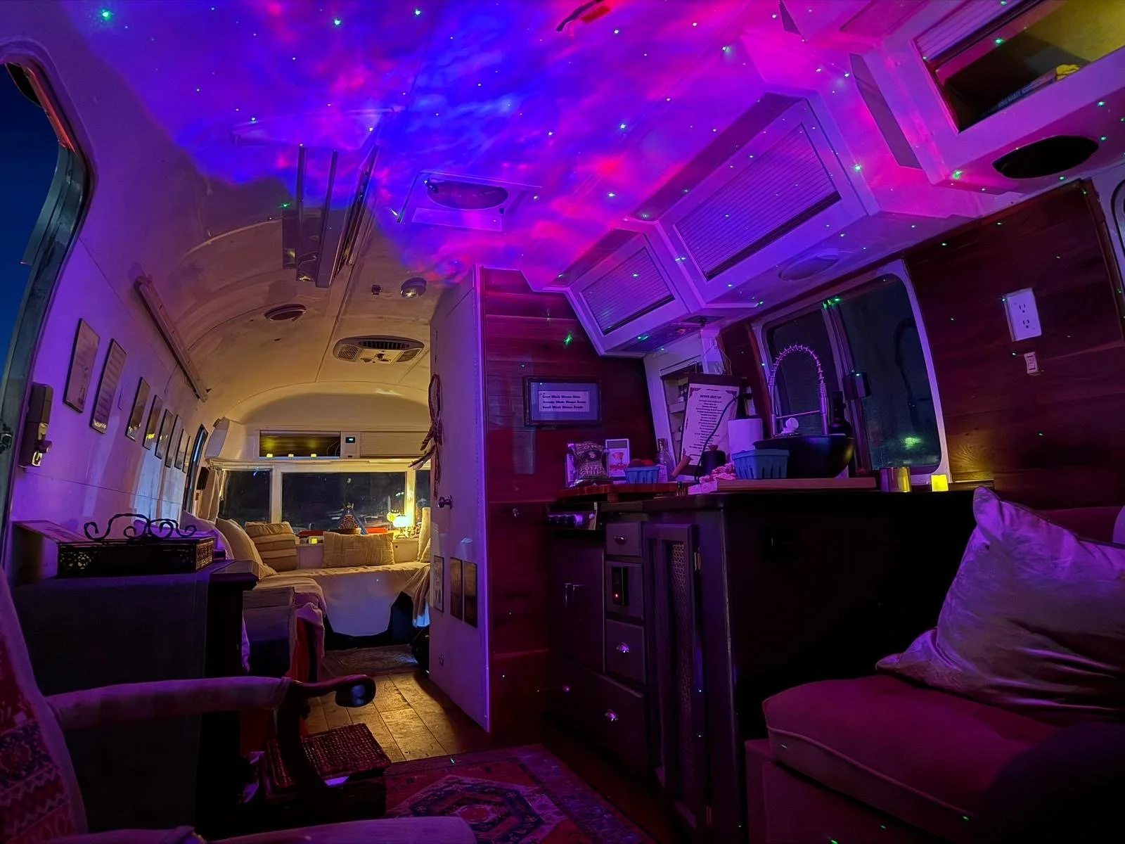 The interior of a cozy Gratitude bus illuminated with colorful LED lights, featuring a small kitchenette with a black sink, a seating area with pillows, and a window with a view of the night outside.