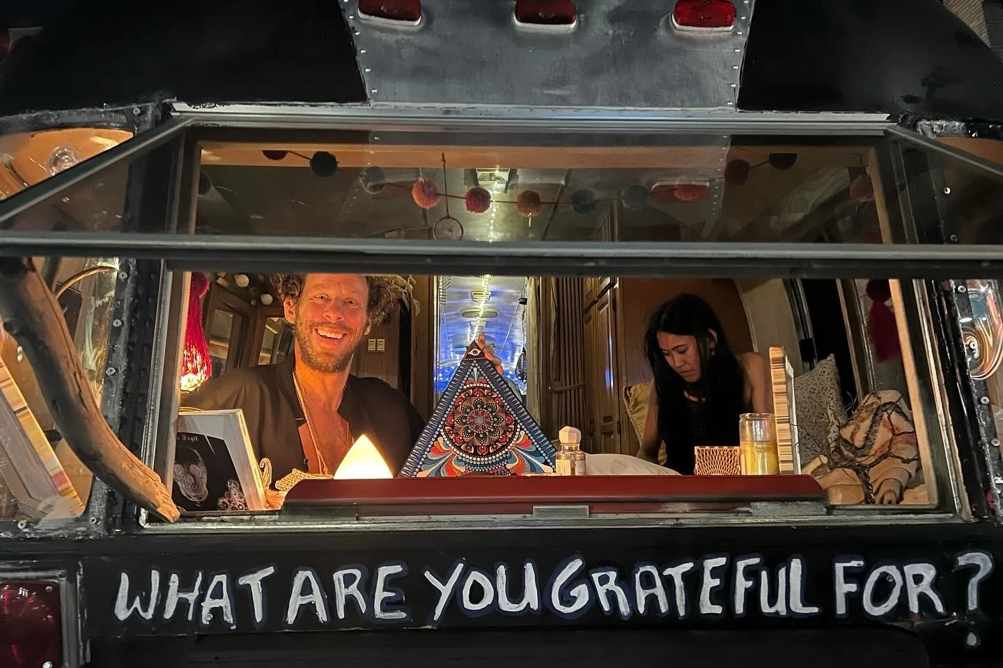 A decorated food truck with the phrase 'WHAT "WHAT ARE YOU GRATEFUL FOR?' written on the back. Doug Akin smiles at the camera, and a woman is looking down, engaged in an activity. The interior has warm lighting and various items, including a colorful