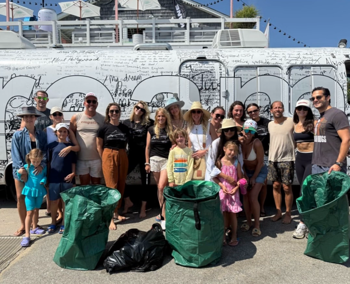 Group of people standing outdoors in front of a large graffiti-covered vehicle, with several large green recycling bags and black garbage bags placed on the ground in front.