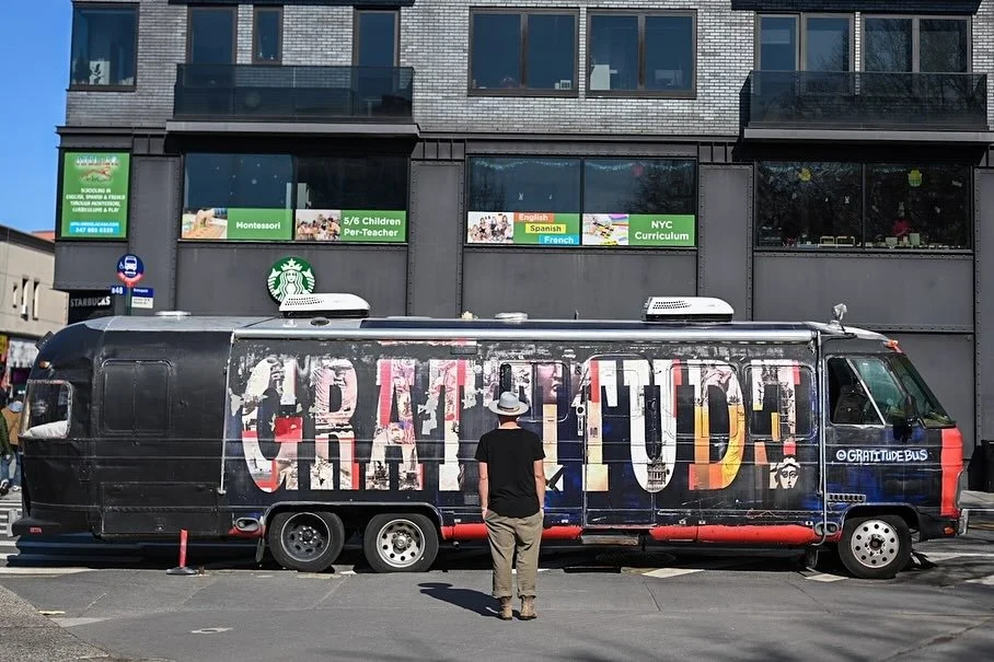 A large bus with the word 'GRATITUDE' painted on its side in colorful, splattered letters. A man wearing a hat stands in front of the bus in an urban area, with a building and windows in the background.