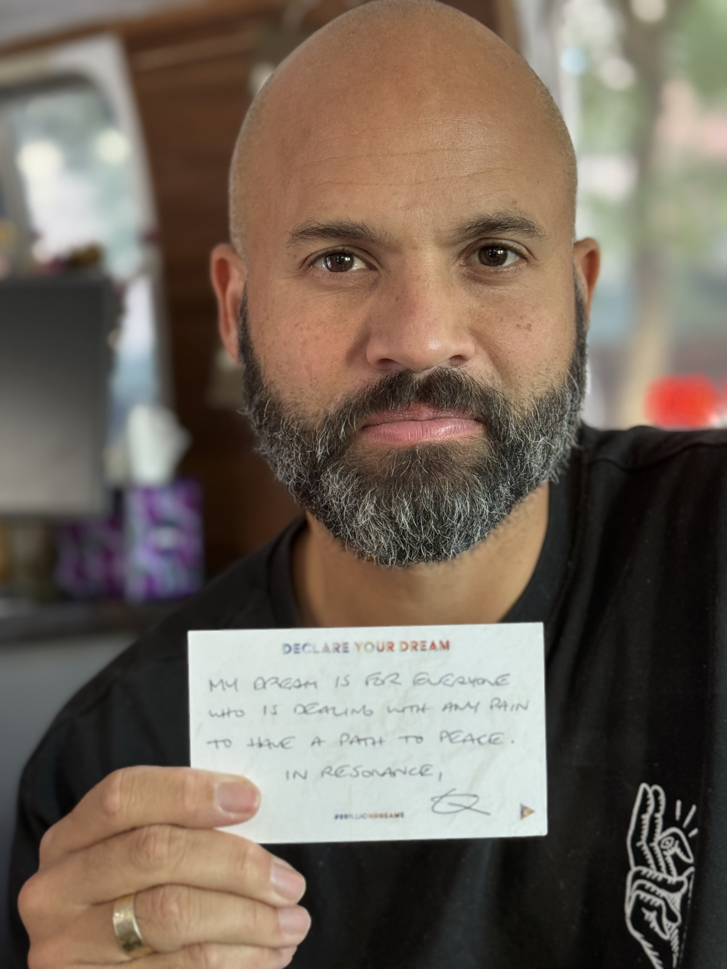 A man with a beard and bald head holding a handwritten note, with a blurred background that appears to be a cozy indoor setting.