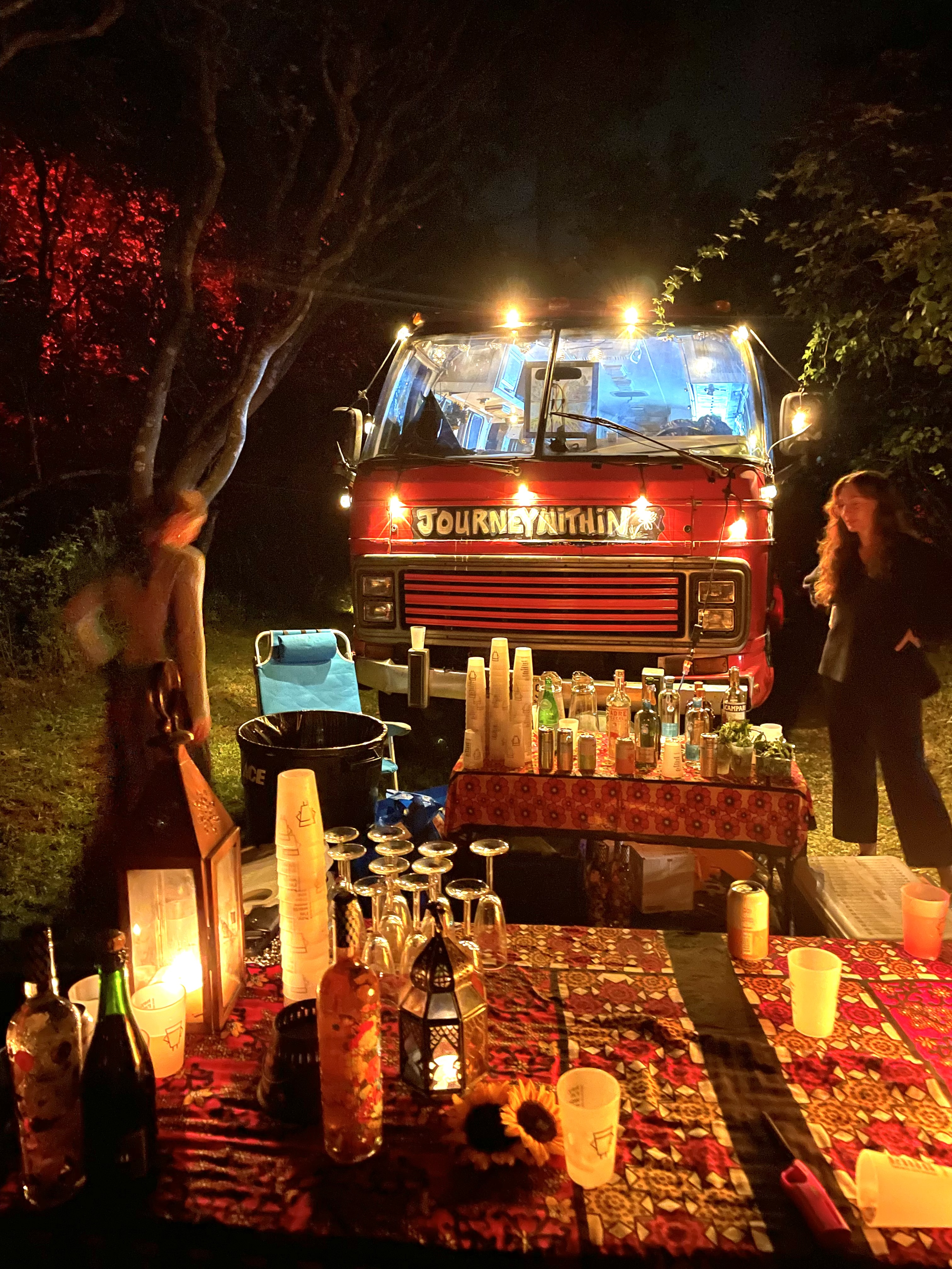 Night scene featuring a decorated food or drink stand with a red truck labeled "Journey Within" in a wooded outdoor setting. The stand is illuminated with string lights, and there are bottles, cups, and decorative items on a patterned tablecloth, wit