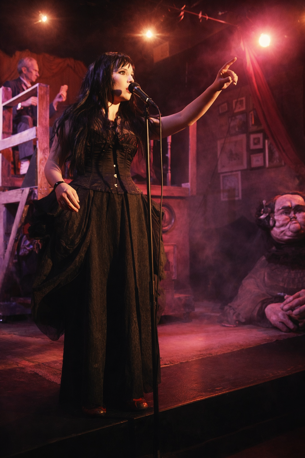 A woman with long dark hair dressed in a black corset and flowing skirt, singing into a microphone on stage, pointing upward, with stage lighting and theatrical set pieces in the background.