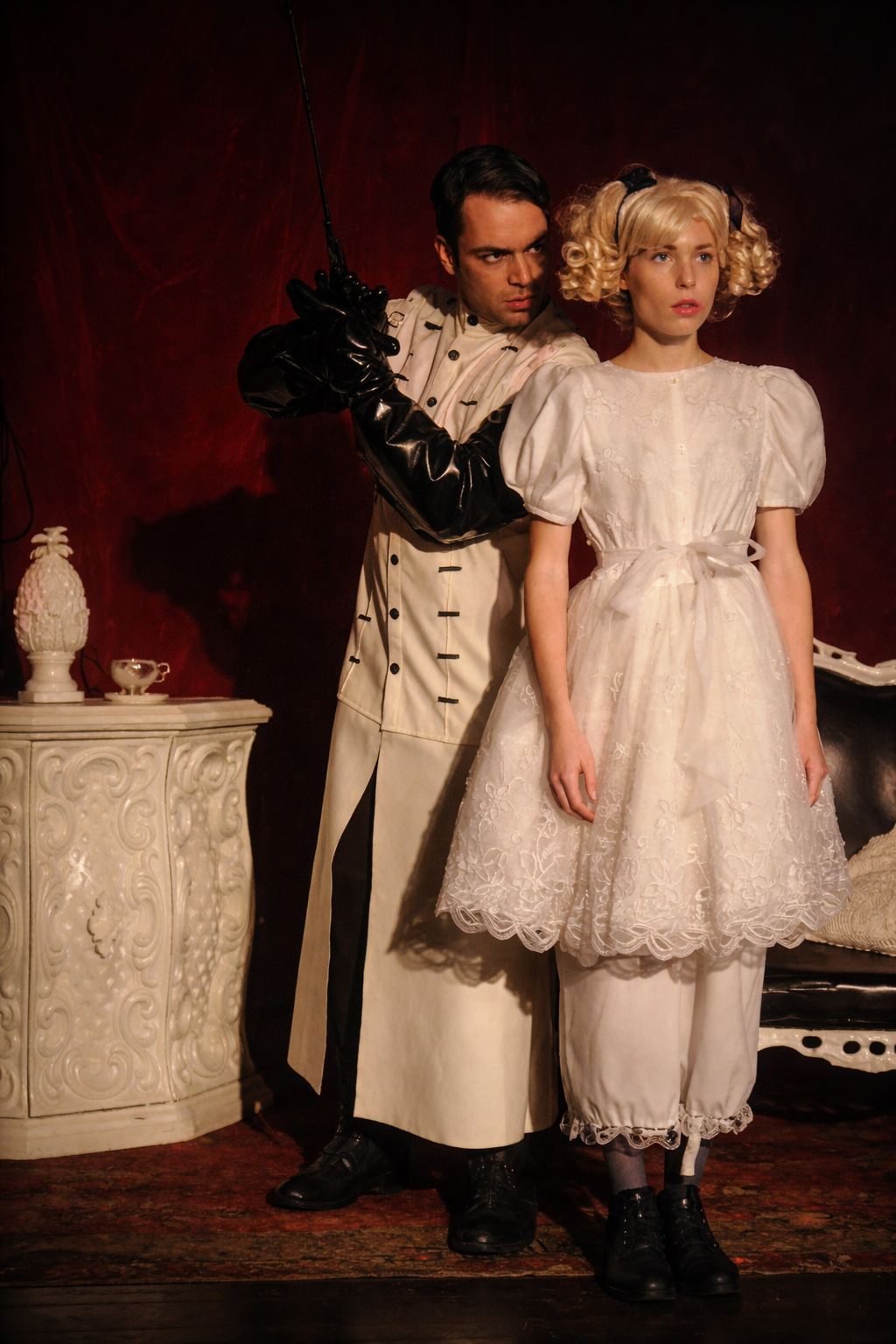 A man wearing black gloves and a white coat with black accents holds a sword behind a young woman in a vintage white dress with puffed sleeves and lace, against a dark red backdrop.