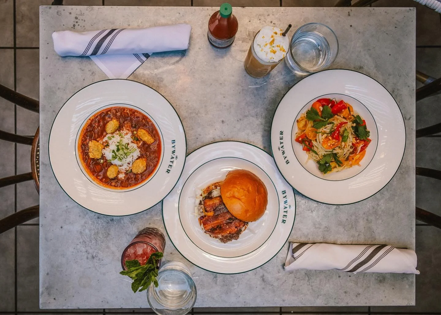 THE NEW STARS OF THE SHOW Summer menu in full bloom we give an ovation three of our favorites. Fresh corn and fried okra give our Tomato gumbo that Louisiana feel. A double smash patty Bywater burger with candied bacon, fried pickles, jalape&ntilde;o