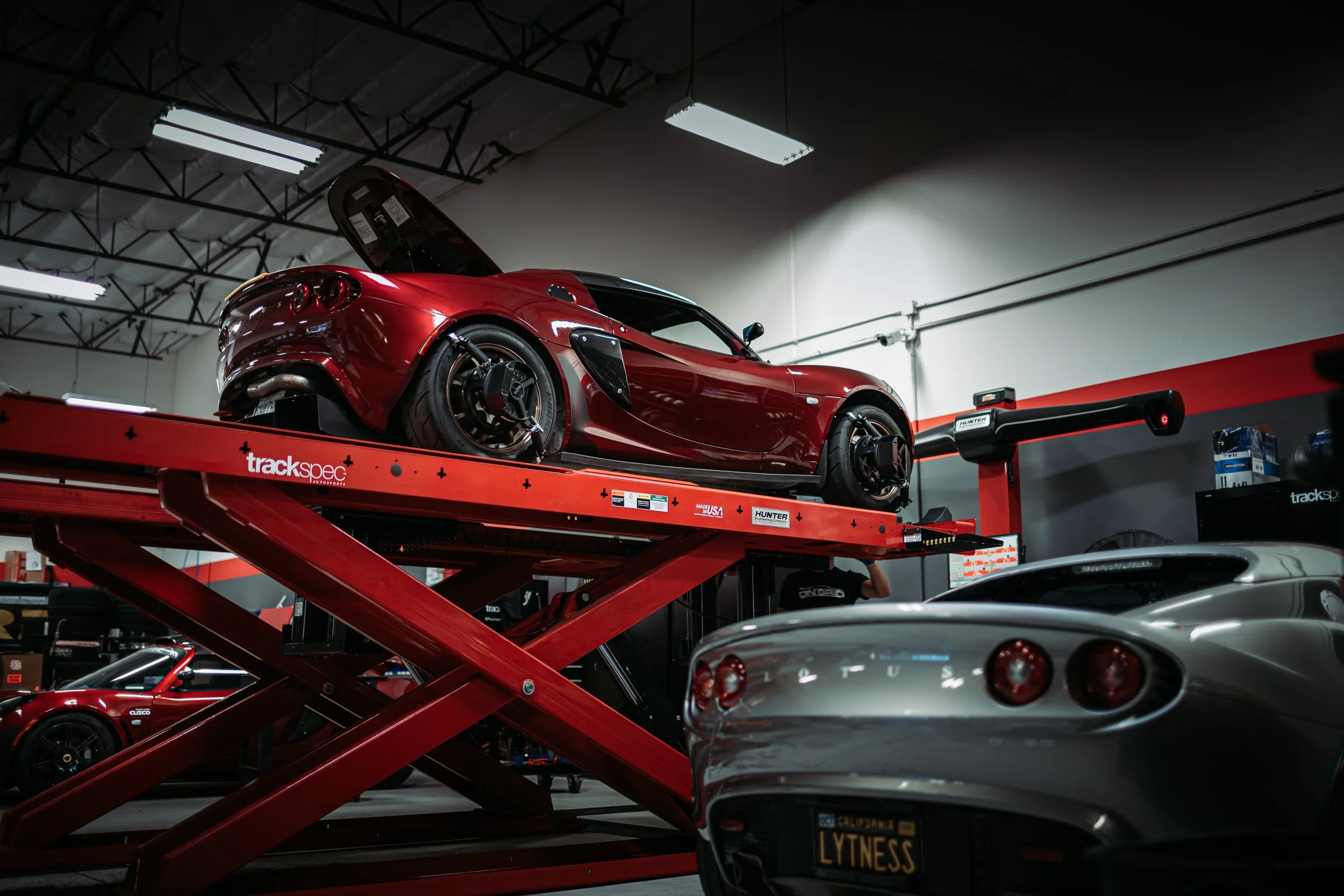 Red sports car on a hydraulic lift in a garage with a silver sports car in the foreground.