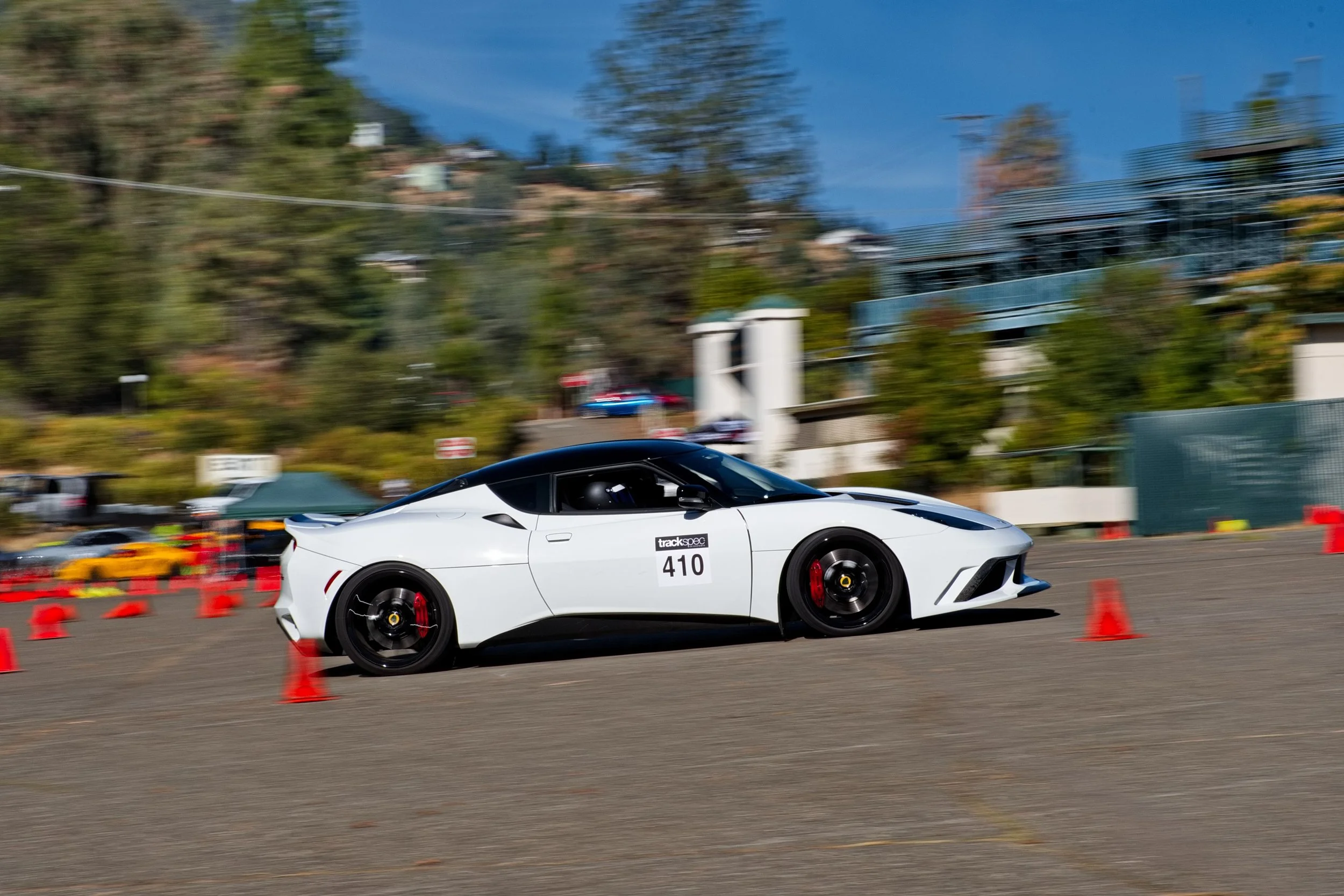 A white sports car navigating a slalom course marked by orange cones at a track event, with a backdrop of trees and buildings.