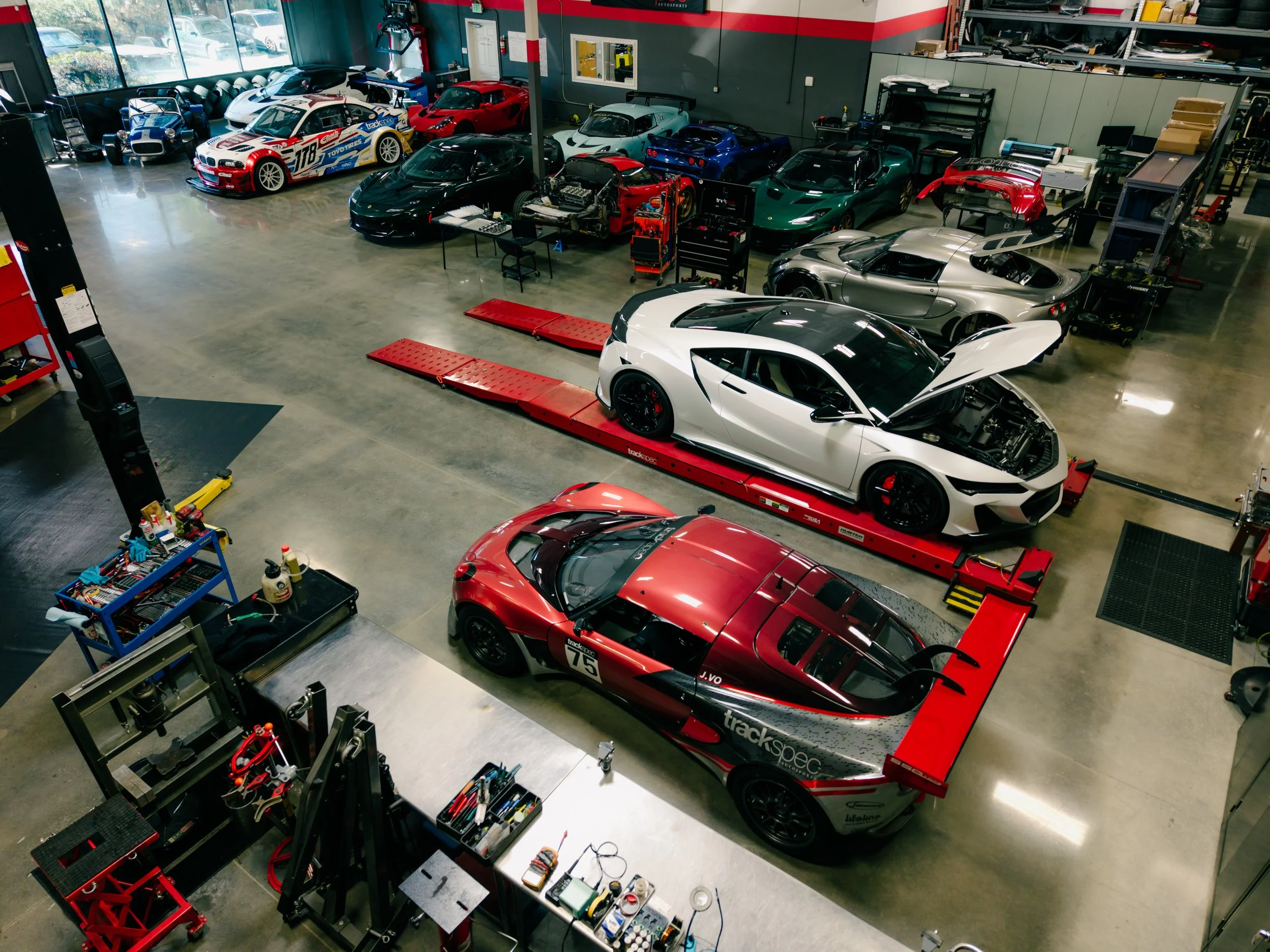 A spacious garage with several high-performance sports cars, including a white Lamborghini with its hood open on a red lift and a red race car labeled number 75, among other luxury and race cars parked inside. There are workbenches and tools along th