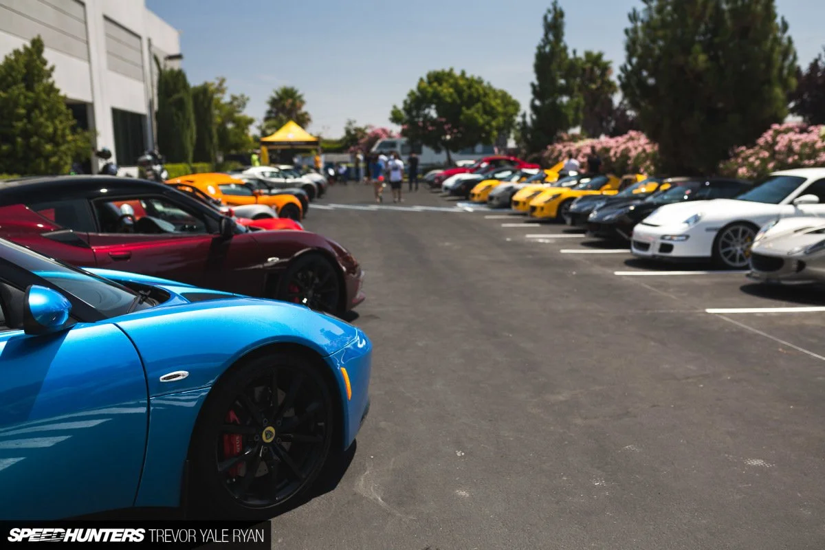 Various luxury sports cars parked in a lot during a car event, with people walking around and blooming trees in the background.