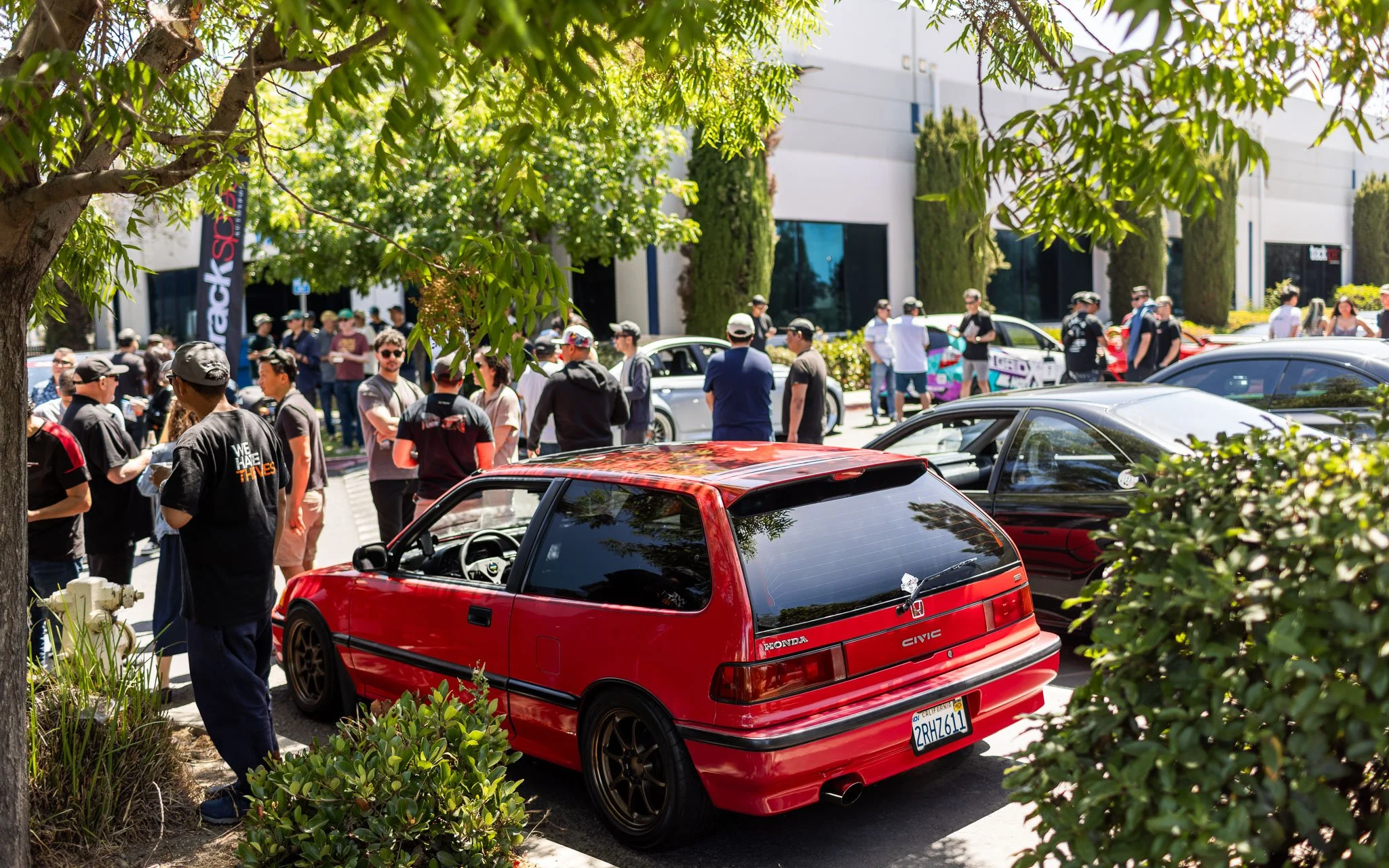 Red Honda Civic hatchback parked in a crowded outdoor event with people socializing and cars on display in the background, shaded by a tree.