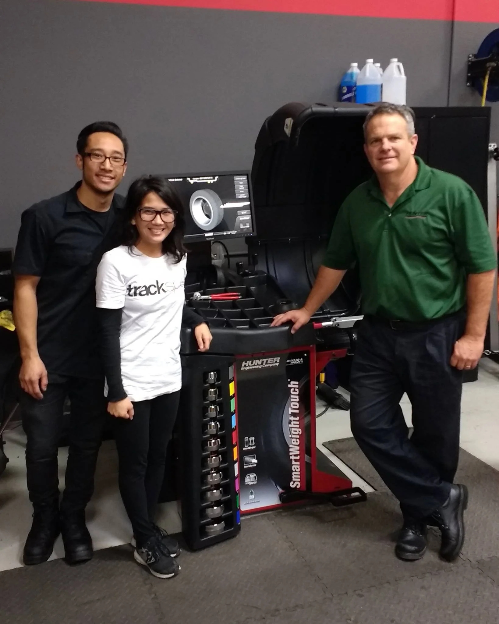 Three people standing next to a tire balancing machine inside a garage or workshop. The man on the right is dressed in a green shirt, and the two individuals on the left are smiling, with one wearing a white t-shirt. The background shows bottles and equipment.