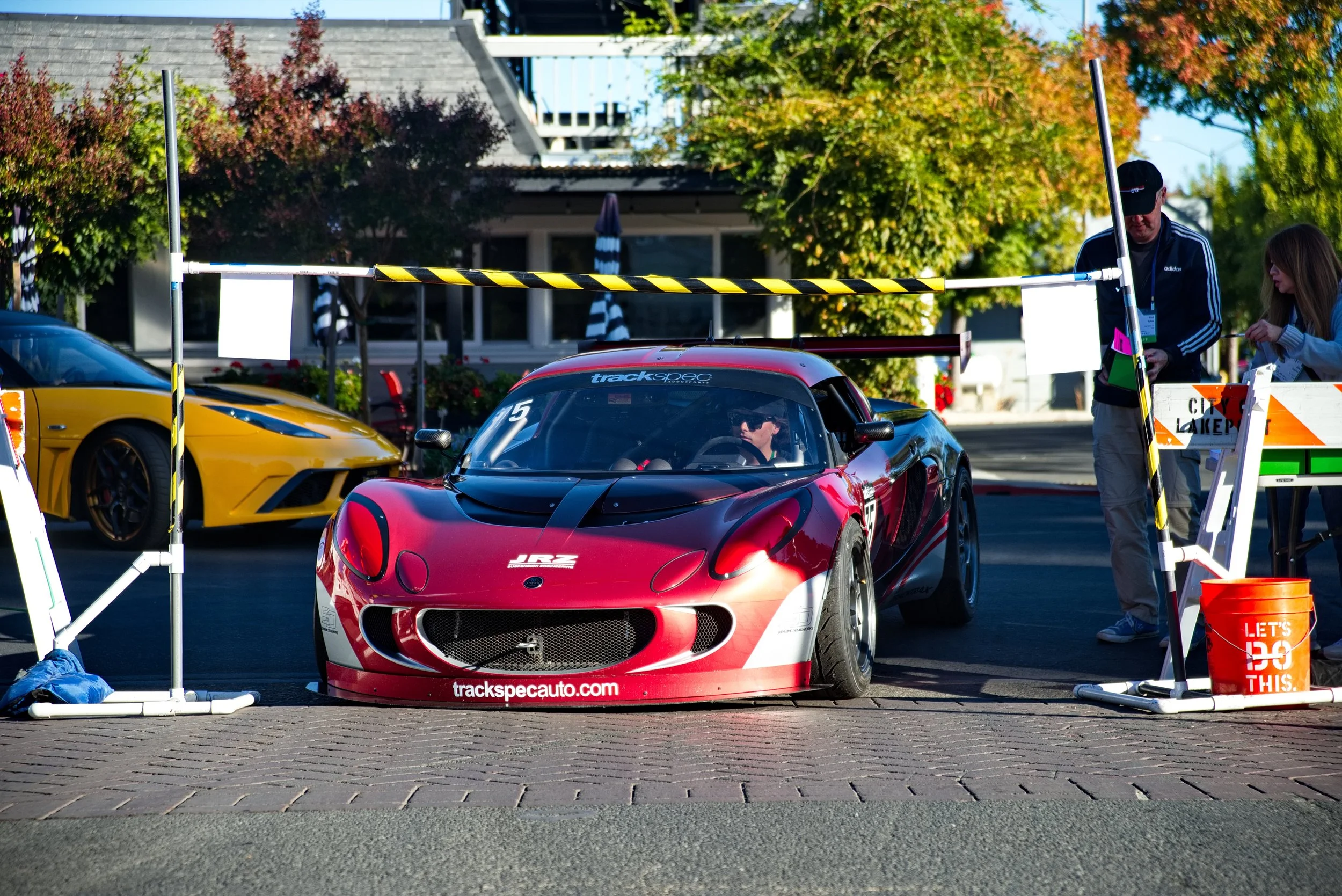 Red and black race car parked behind a starting gate at a street racing event, with a yellow vehicle in the background and two people standing nearby.