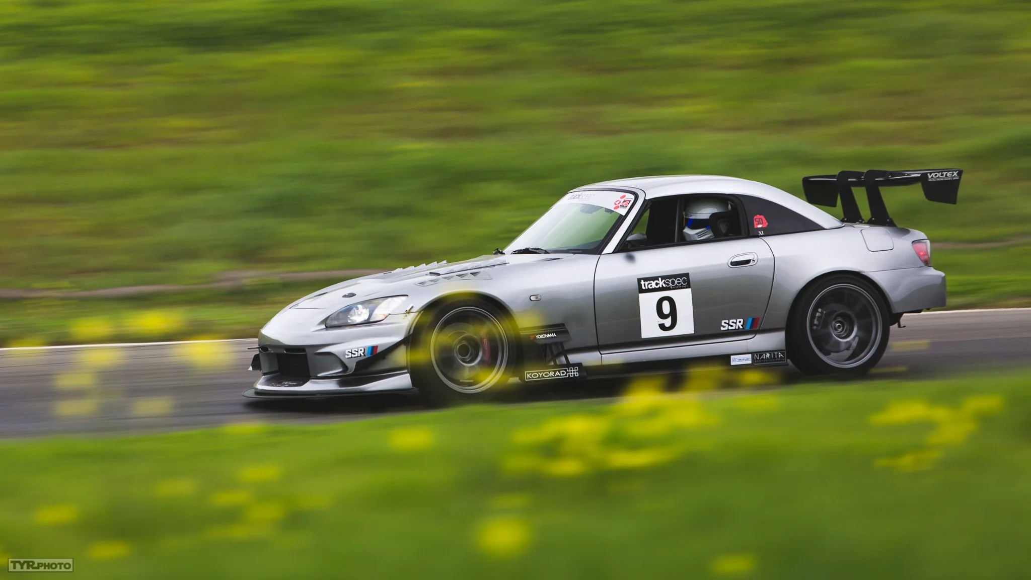 A silver racing car with the number 9 on it driving fast on a track, with a green field and yellow flowers in the foreground.