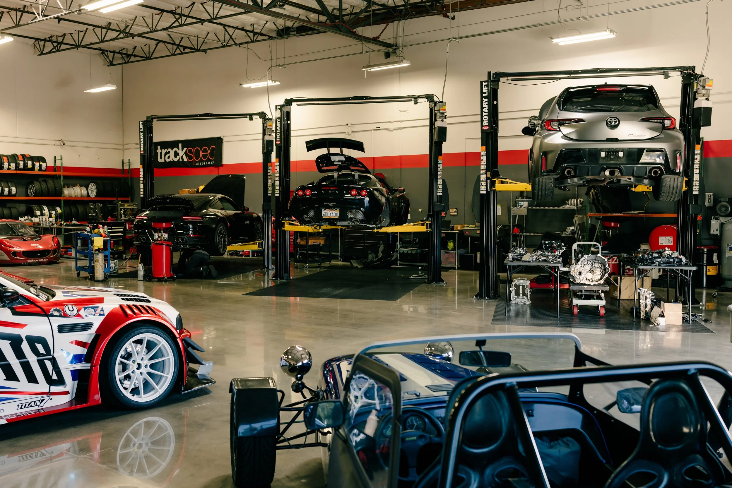 Indoor garage with racing cars on lifts and high-performance car parts arranged on tables.
