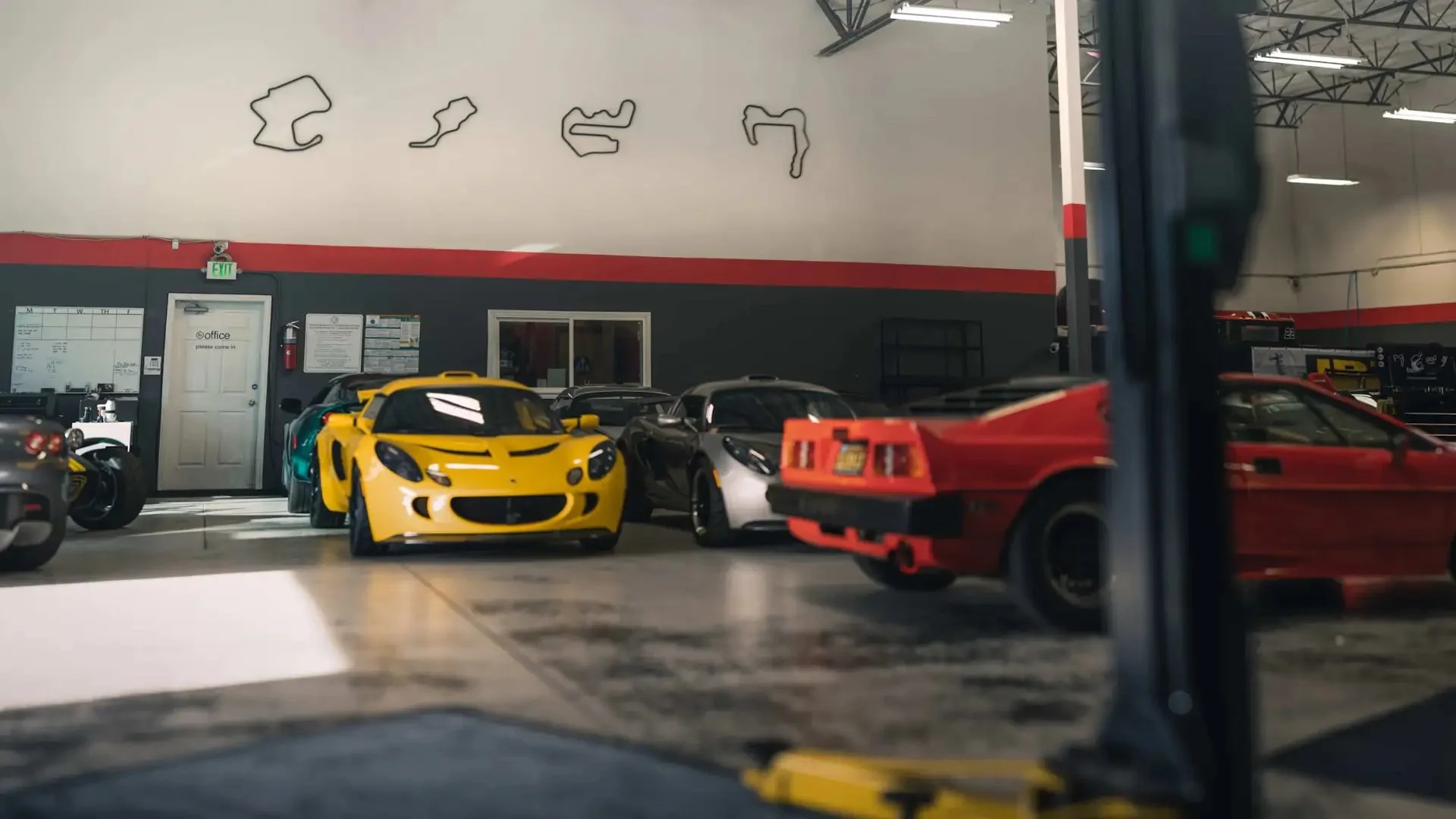 A garage filled with various model cars, including a yellow sports car in the front, a silver and black car behind it, and a red car on the right side. The background features a wall with abstract black line art and an office door.