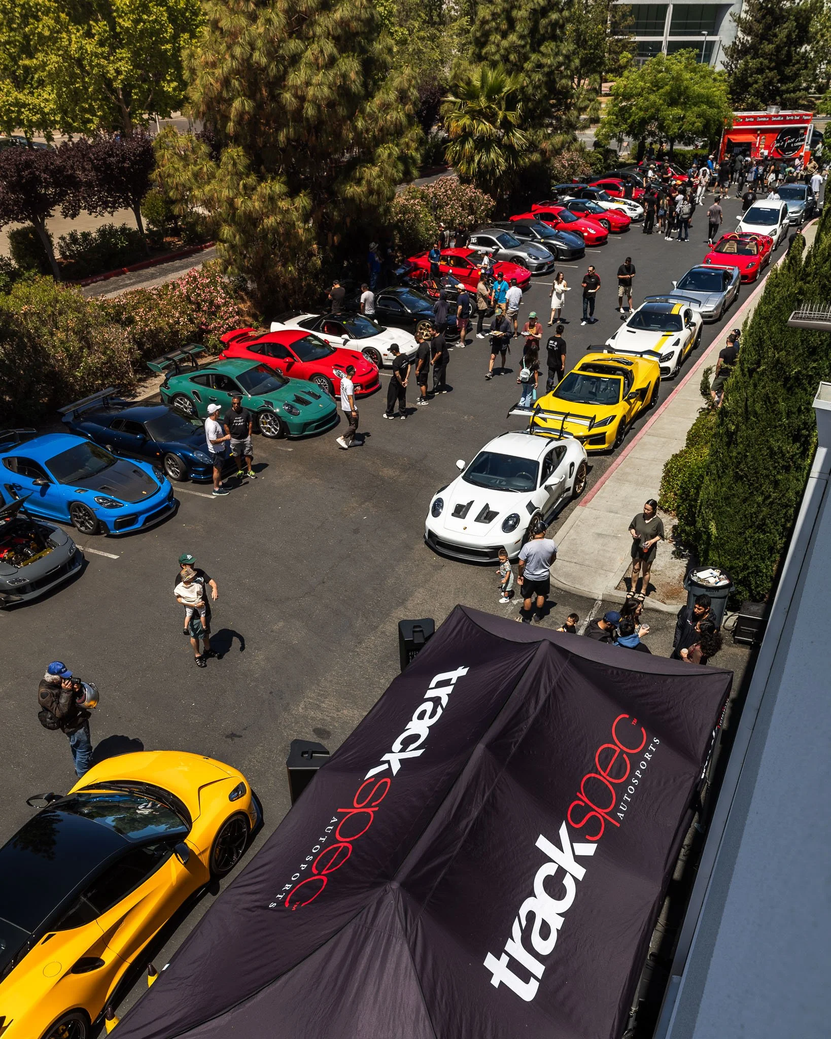 An outdoor car meet-up displaying various high-end and sports cars parked in a lot, with people walking around and viewing the vehicles, view from a high-angle.