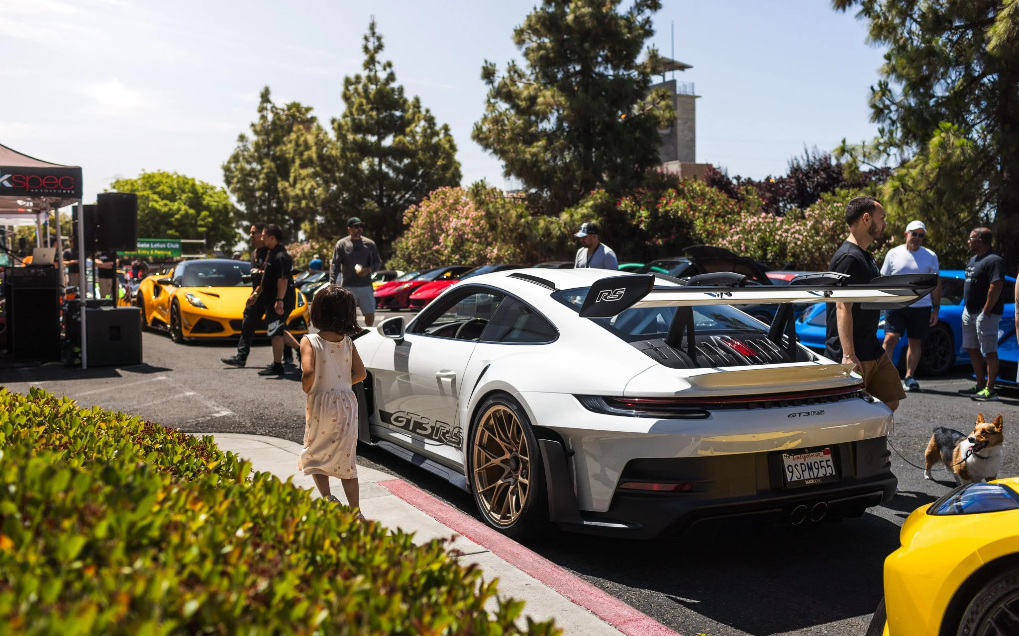 A white Porsche GT3 RS parked at a car show with a large rear wing, surrounded by a group of people and other colorful sports cars in the background.